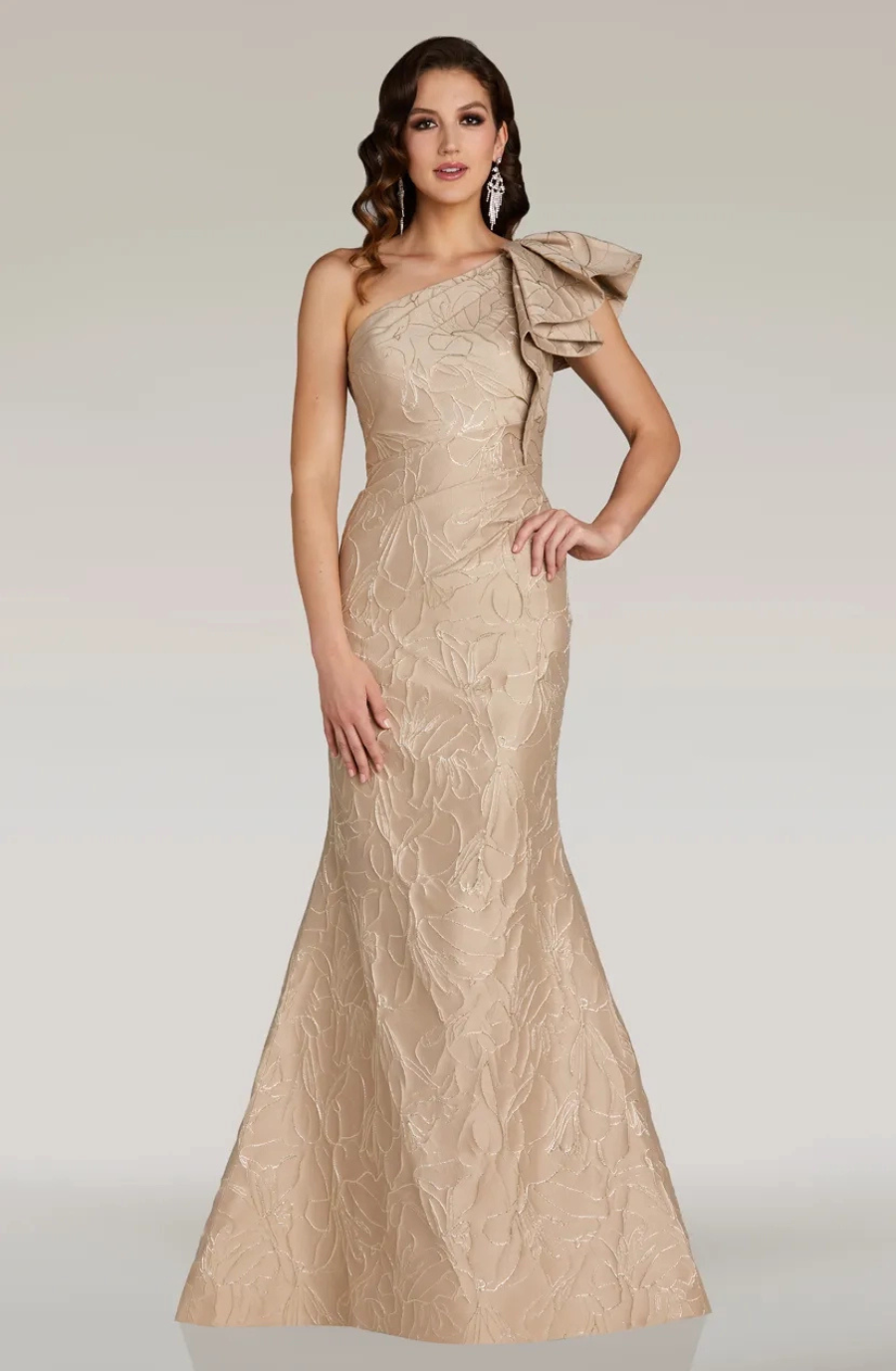 A model wearing a beige, one-shoulder evening gown with a bow detail on the shoulder.