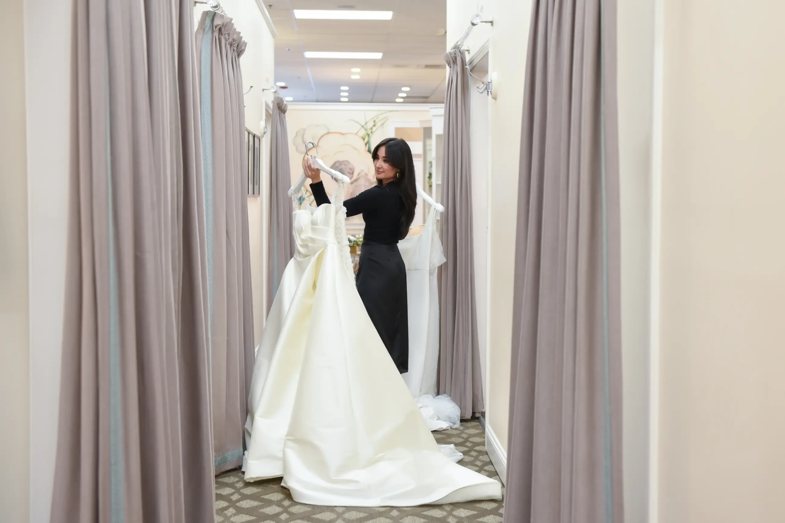 A woman in a black dress stands in a fitting room corridor, smiling as she holds two white wedding dresses on hangers. The scene is elegant and joyful.