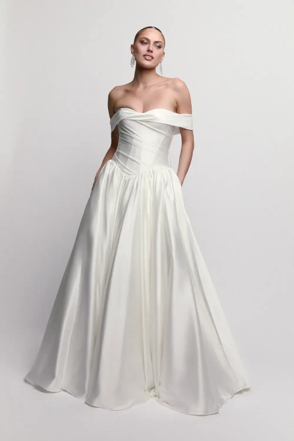 A woman wearing an off-the-shoulder white gown, posing confidently with her hands in the dress pockets.