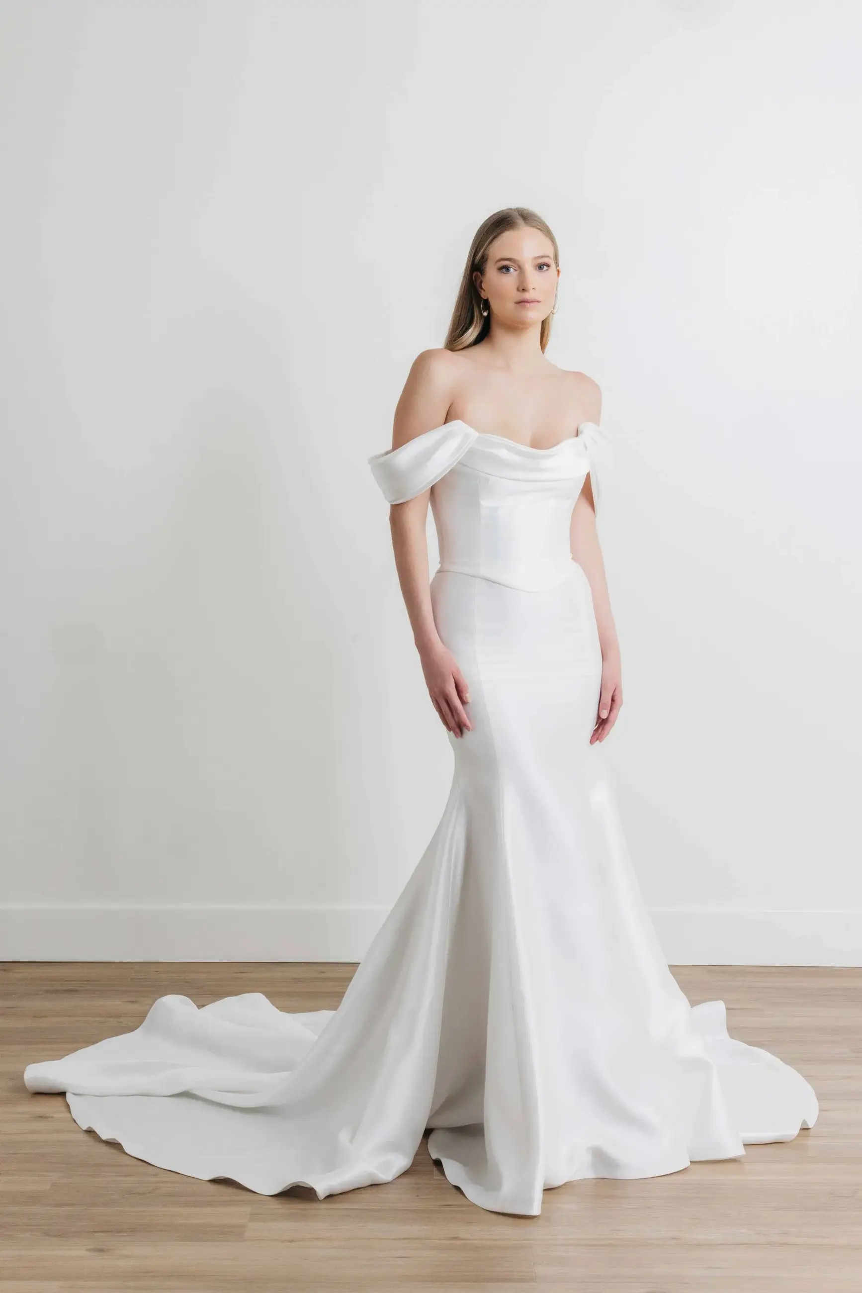 Model is wearing a fit and flare off the shoulder bridal gown with a corset bodice and ruching on the neckline