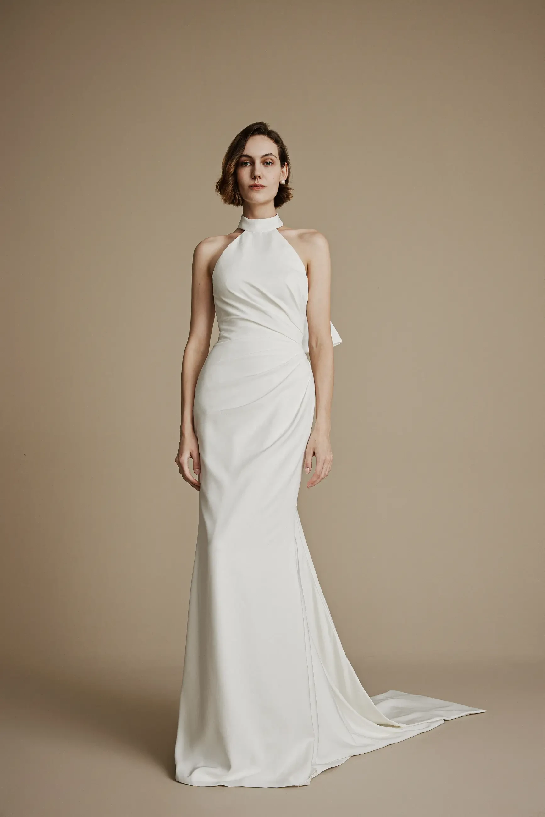 A woman in a white, halter-neck evening gown stands against a beige background. The dress is elegant and form-fitting, with a gentle train, exuding a refined and sophisticated tone.