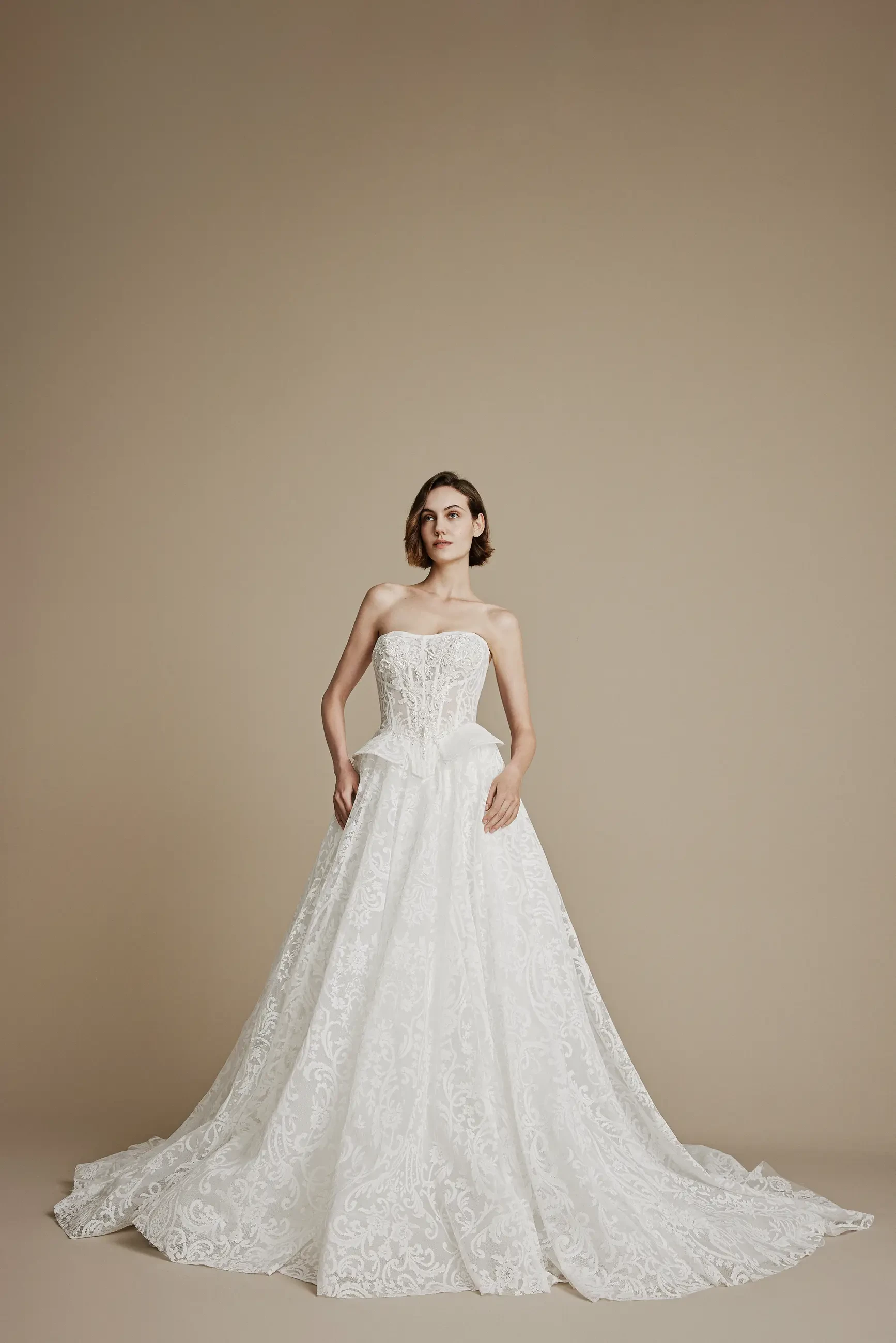 A woman stands against a beige background, wearing an elegant, strapless white lace wedding gown with a fitted bodice and flowing skirt, creating a timeless, sophisticated look.