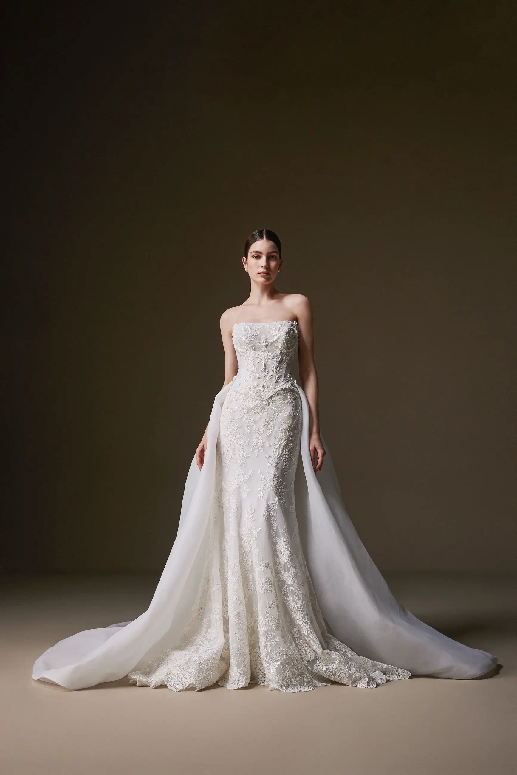A bride in an elegant strapless, embroidered white gown with a long flowing train poses against a dark background, evoking grace and sophistication.