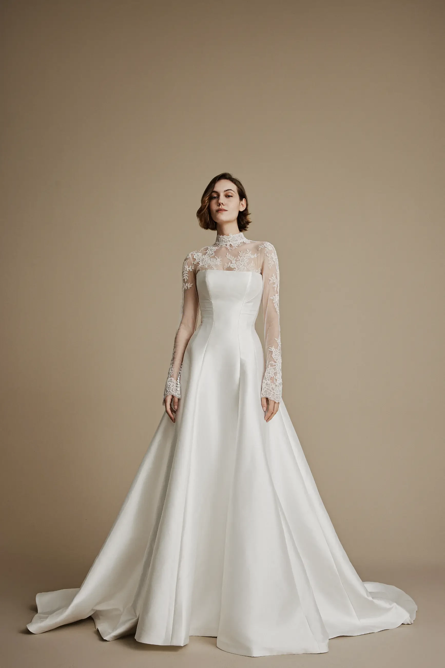 Elegant bride in a white satin gown with lace sleeves and high neckline stands against a neutral background, exuding timeless grace and sophistication.