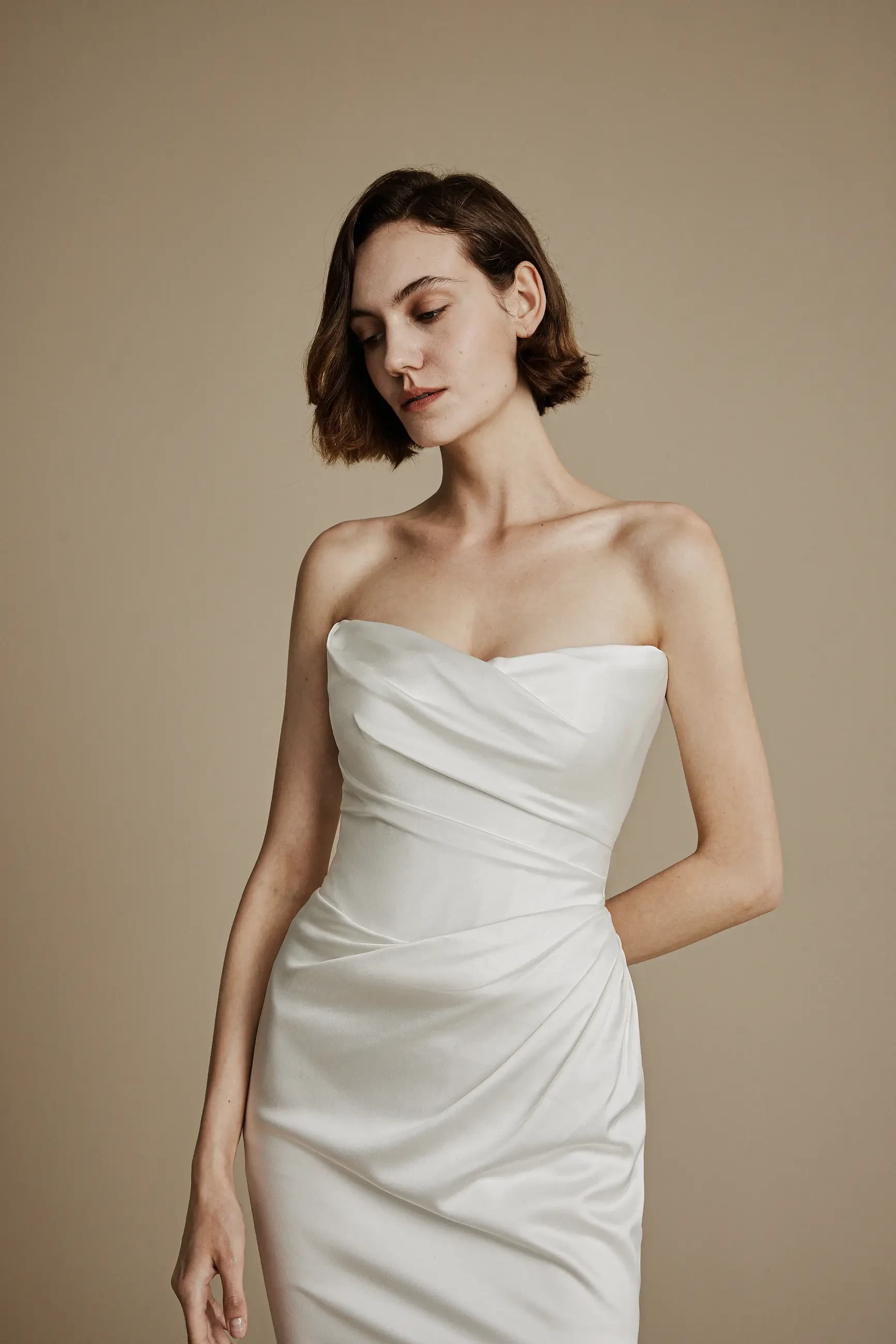 A woman in a strapless, white satin dress poses against a beige background. Her expression is serene, and her short, brown hair is styled simply.