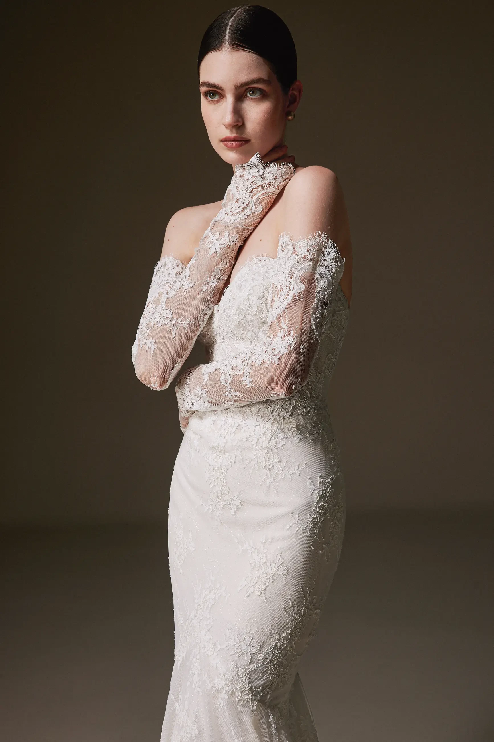 A woman in a strapless, lace bridal gown with matching sheer lace gloves. She has dark hair, styled elegantly, and gazes thoughtfully to the side. The background is softly lit, creating a serene and sophisticated mood.