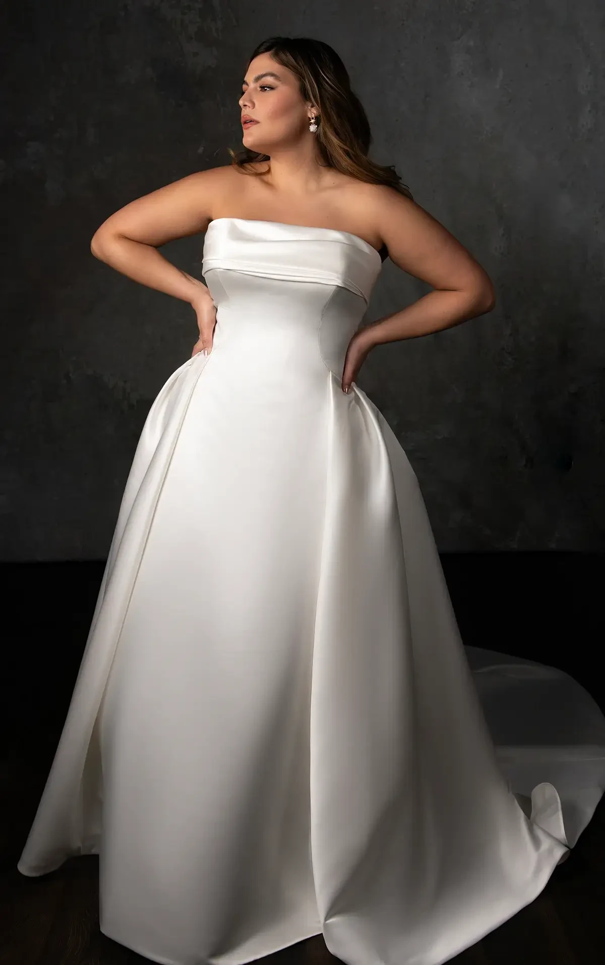 Model wears a strapless satin ball gown with a pleated skirt and fabric-covered buttons down the back.