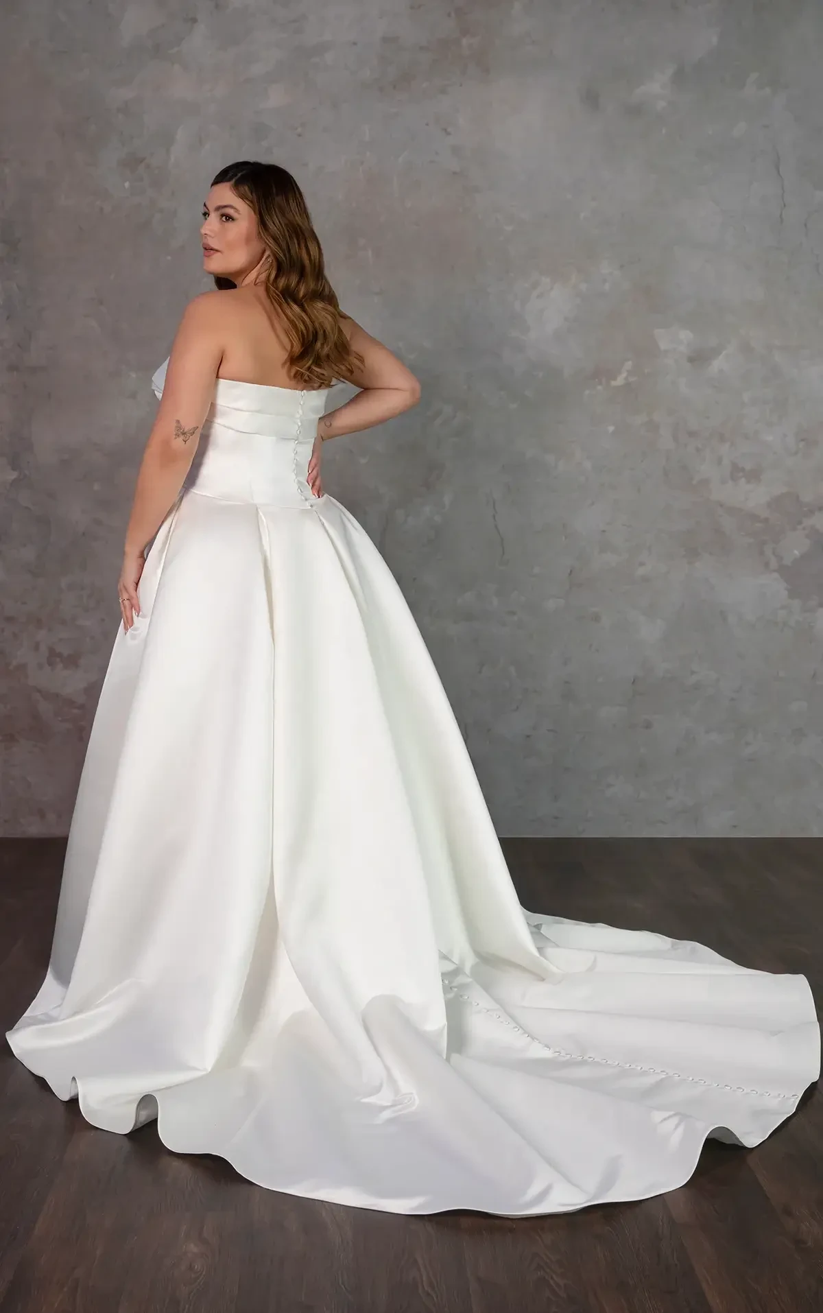 Model wears a strapless satin ball gown with a pleated skirt and fabric-covered buttons down the back.