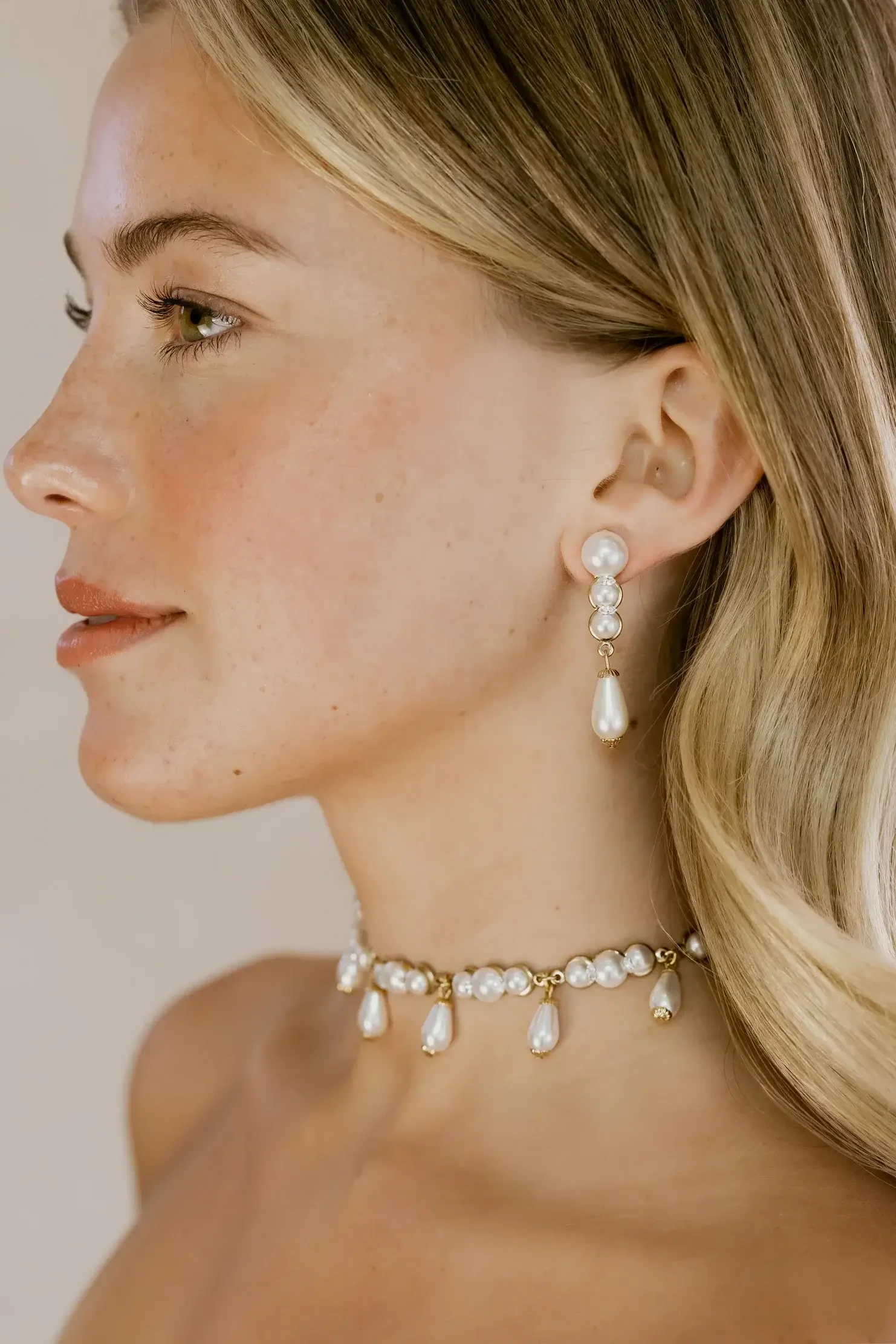 A profile view of a woman with long, wavy blonde hair wearing elegant pearl earrings and a matching pearl necklace.
