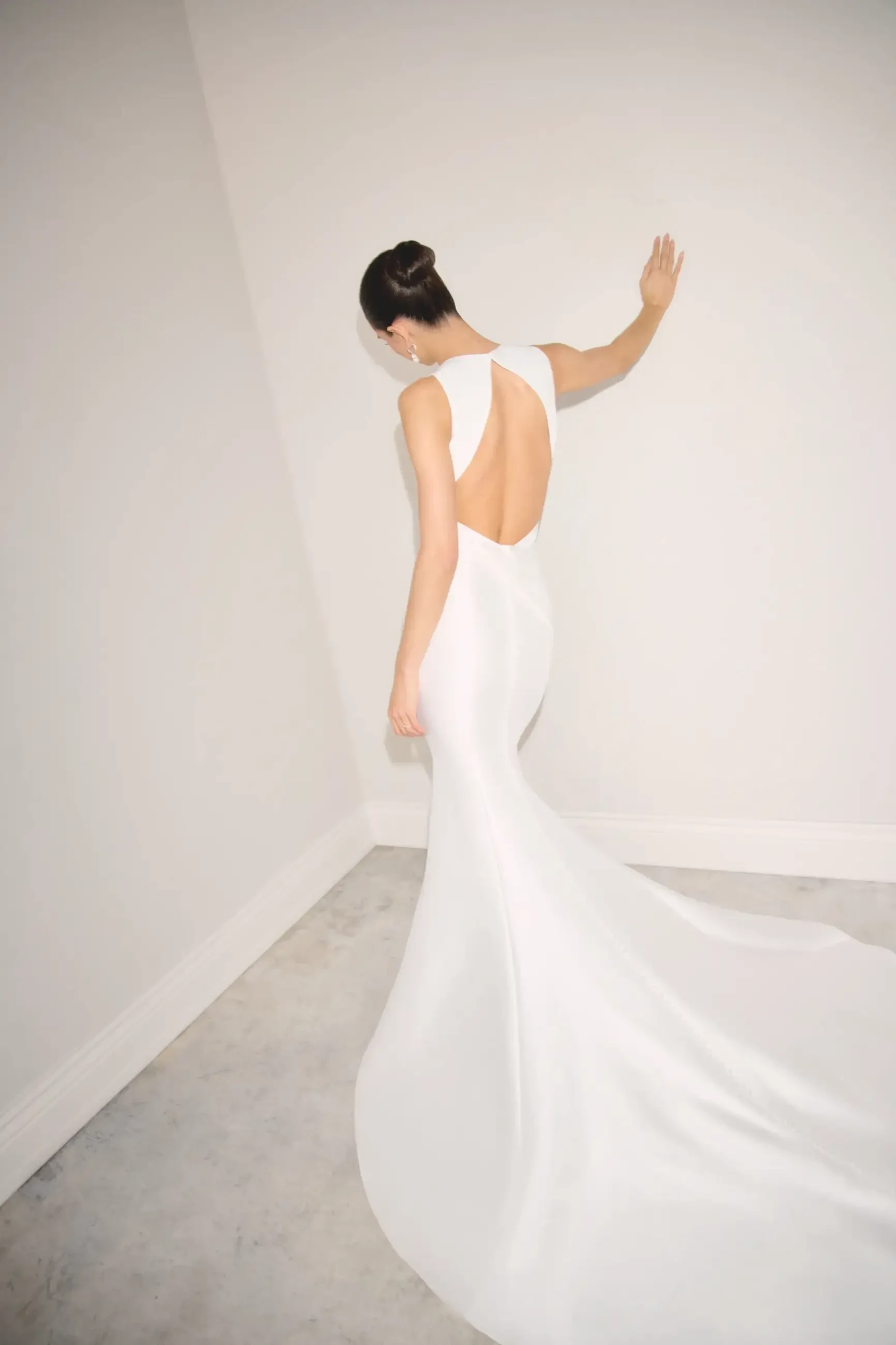 A woman in a sleek, white gown with an open back poses in a minimalist corner, one hand on the wall. The setting is elegant and serene.