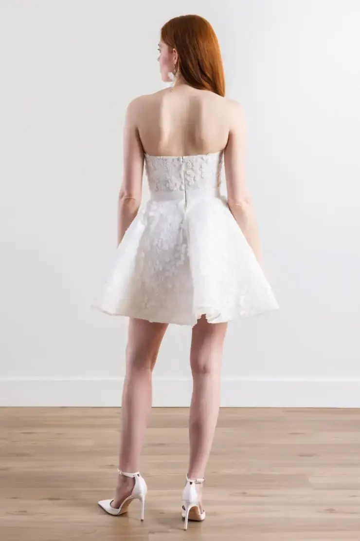 Model is wearing a strapless mini bridal dress featuring a corset bodice, flouncy layered skirt, and floral lace details with a dotted net overlay
