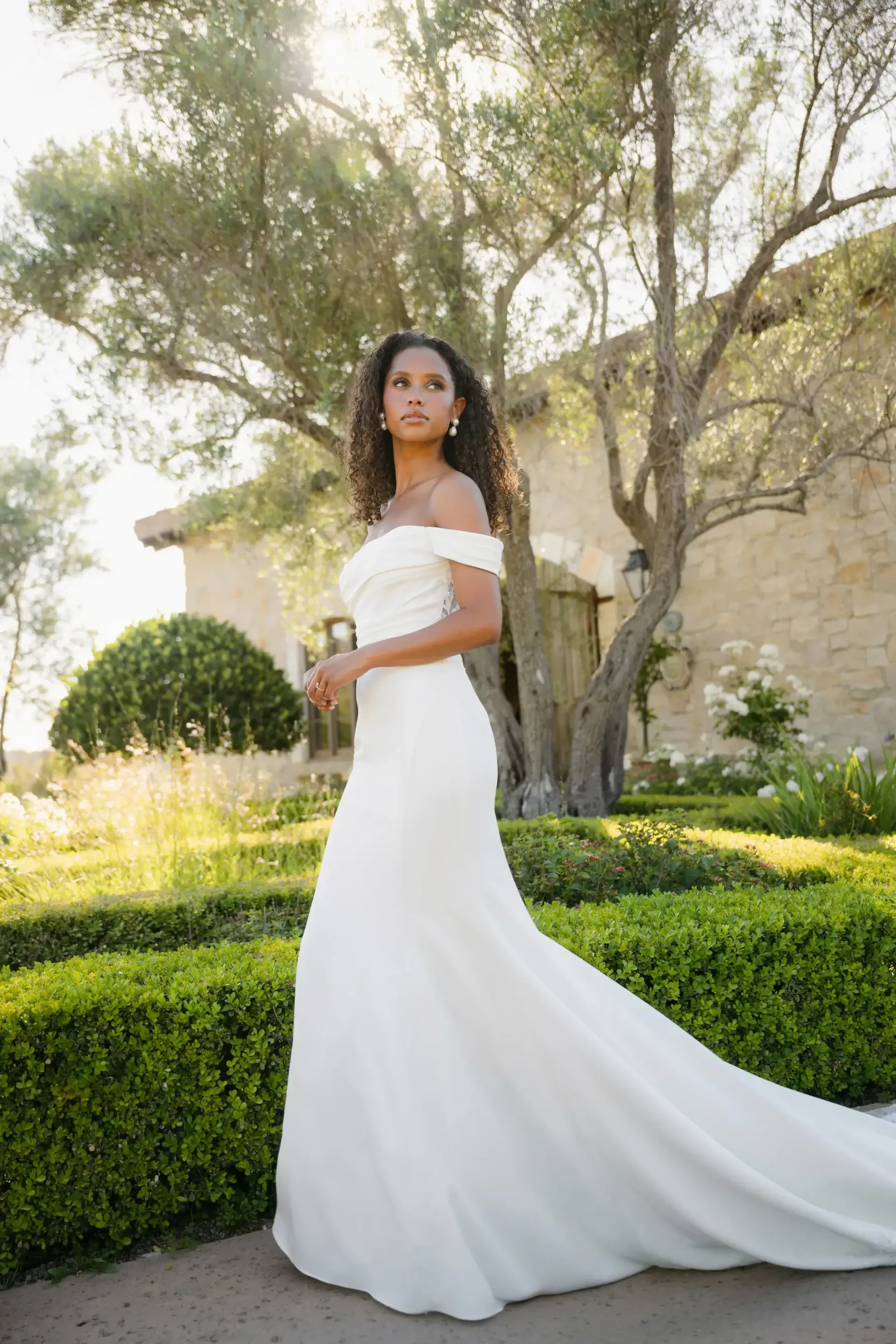 A woman in a flowing white dress stands in a sunlit garden, surrounded by olive trees and a stone building, exuding elegance and tranquility.