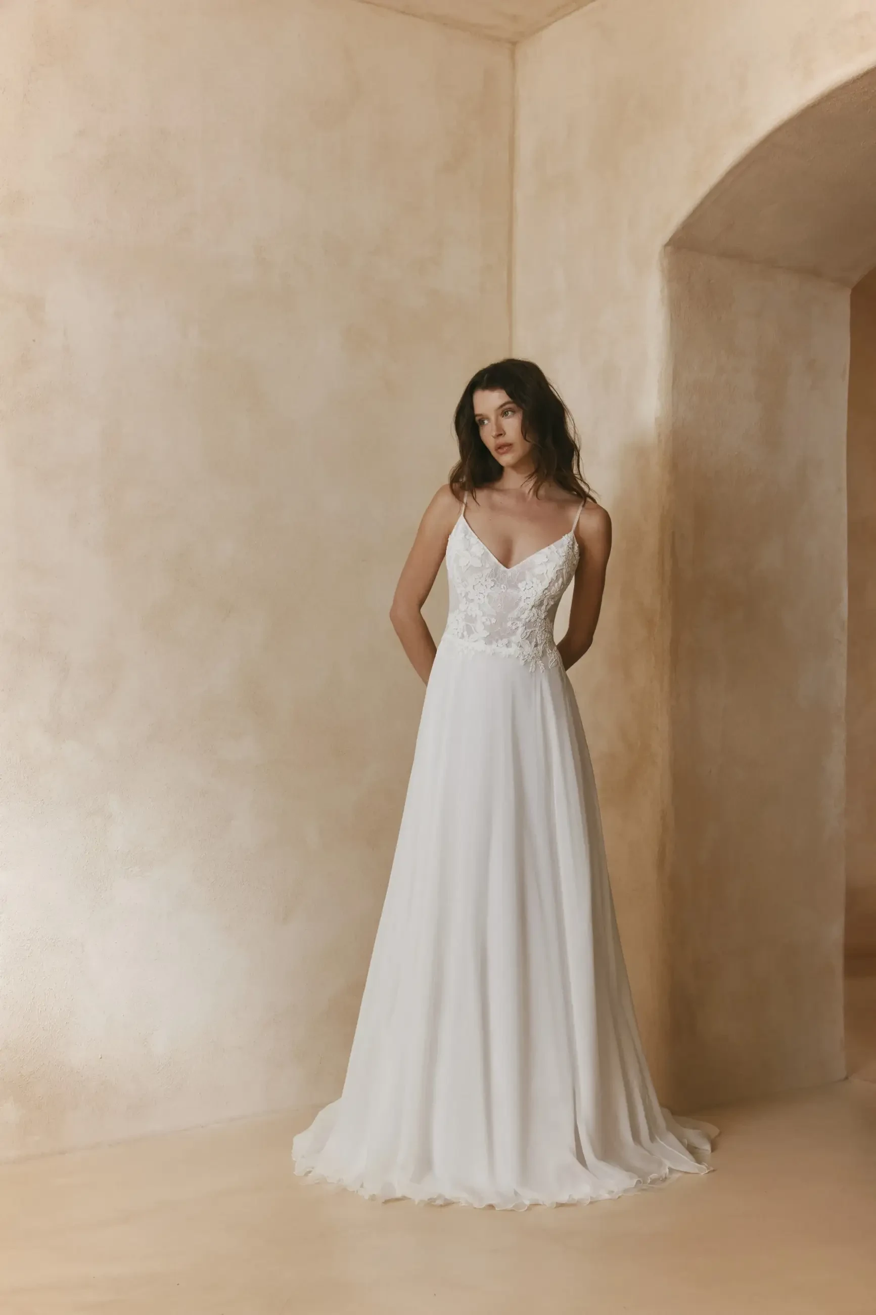 A model wearing a white wedding dress with a delicate bodice, standing against a neutral-toned backdrop.