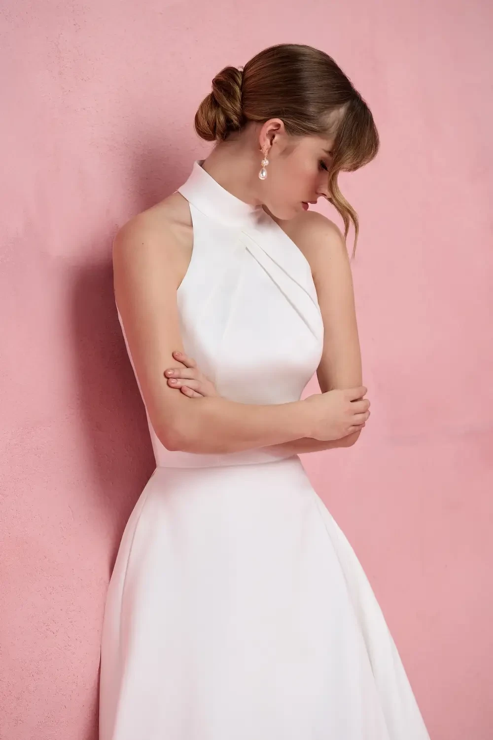 A woman in a white dress with a halter neck, standing against a pink background, with her arms crossed and a thoughtful expression.