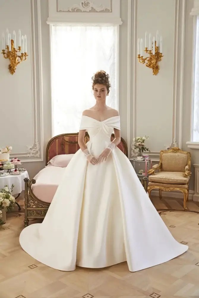 Model is wearing an off the shoulder wedding dress with an overskirt and minimalist sleek fabric