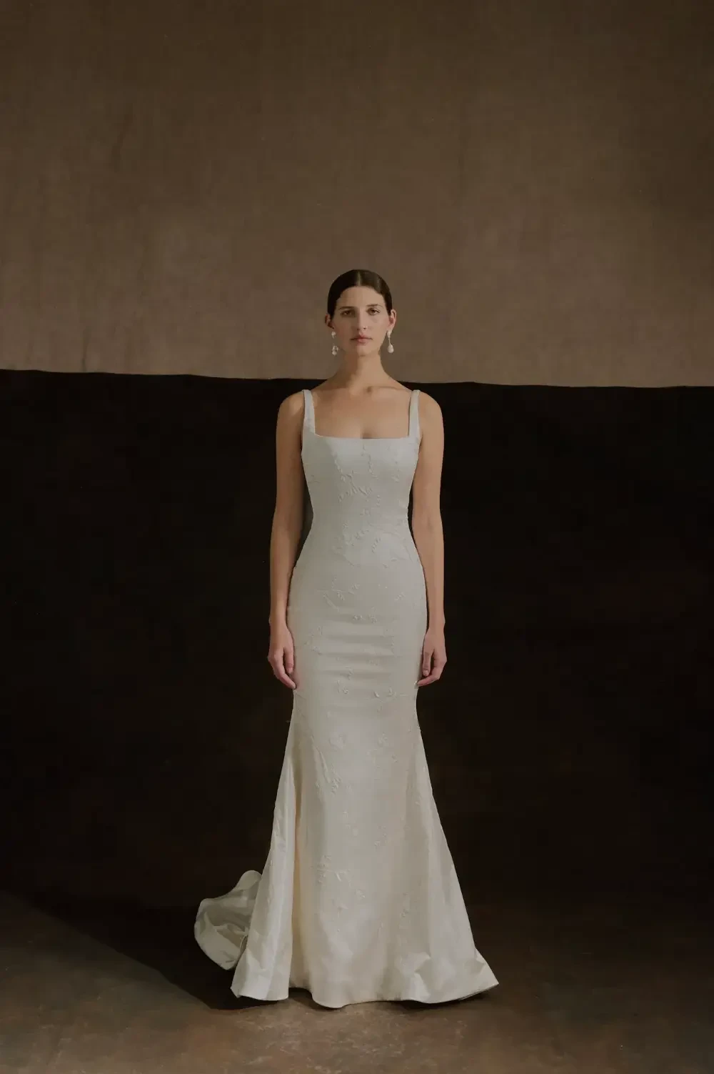 A model wearing a fitted white wedding dress with a square neckline, standing against a neutral backdrop.