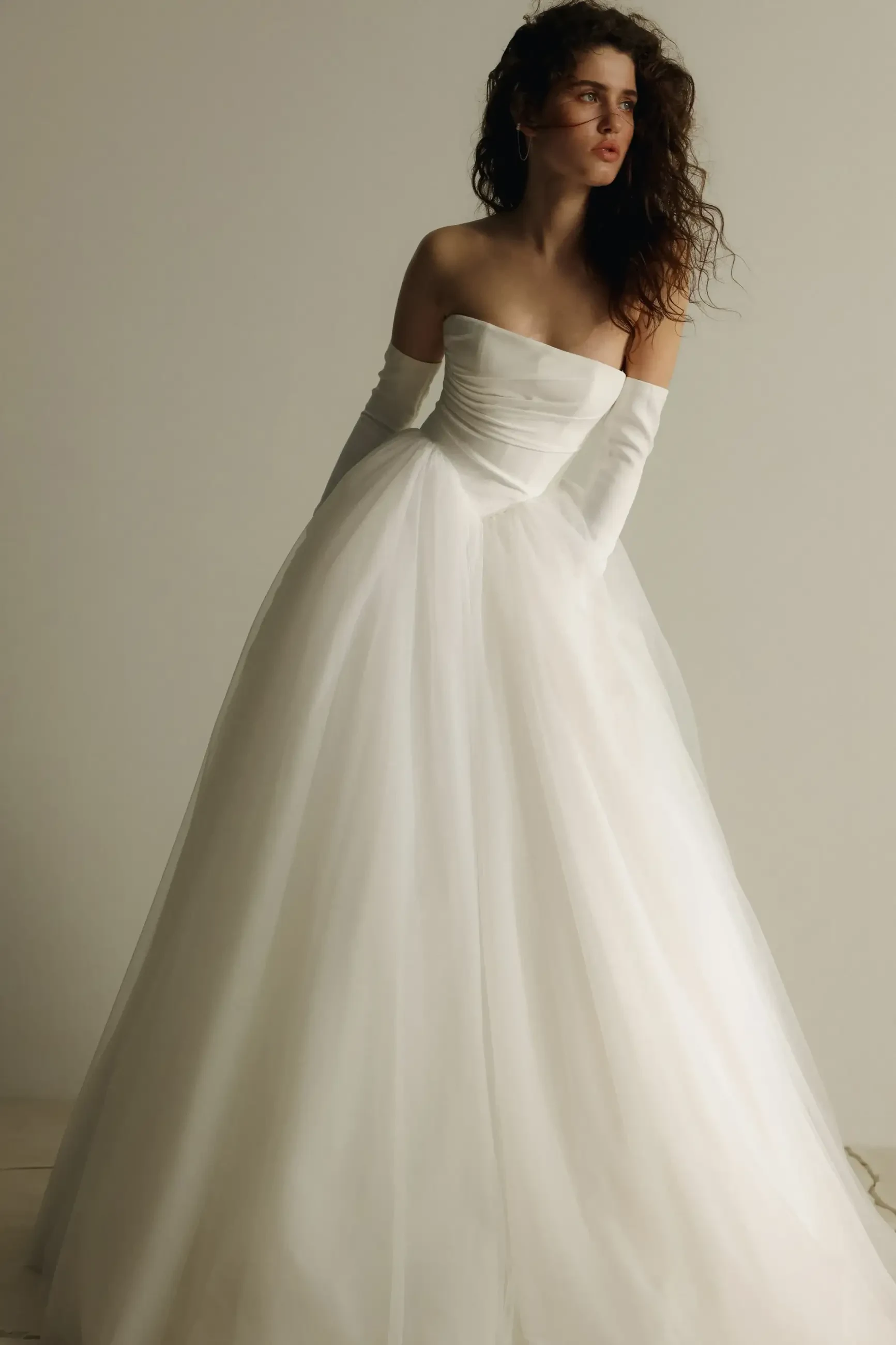 A woman wearing a strapless white wedding gown with a full skirt, standing against a neutral background.
