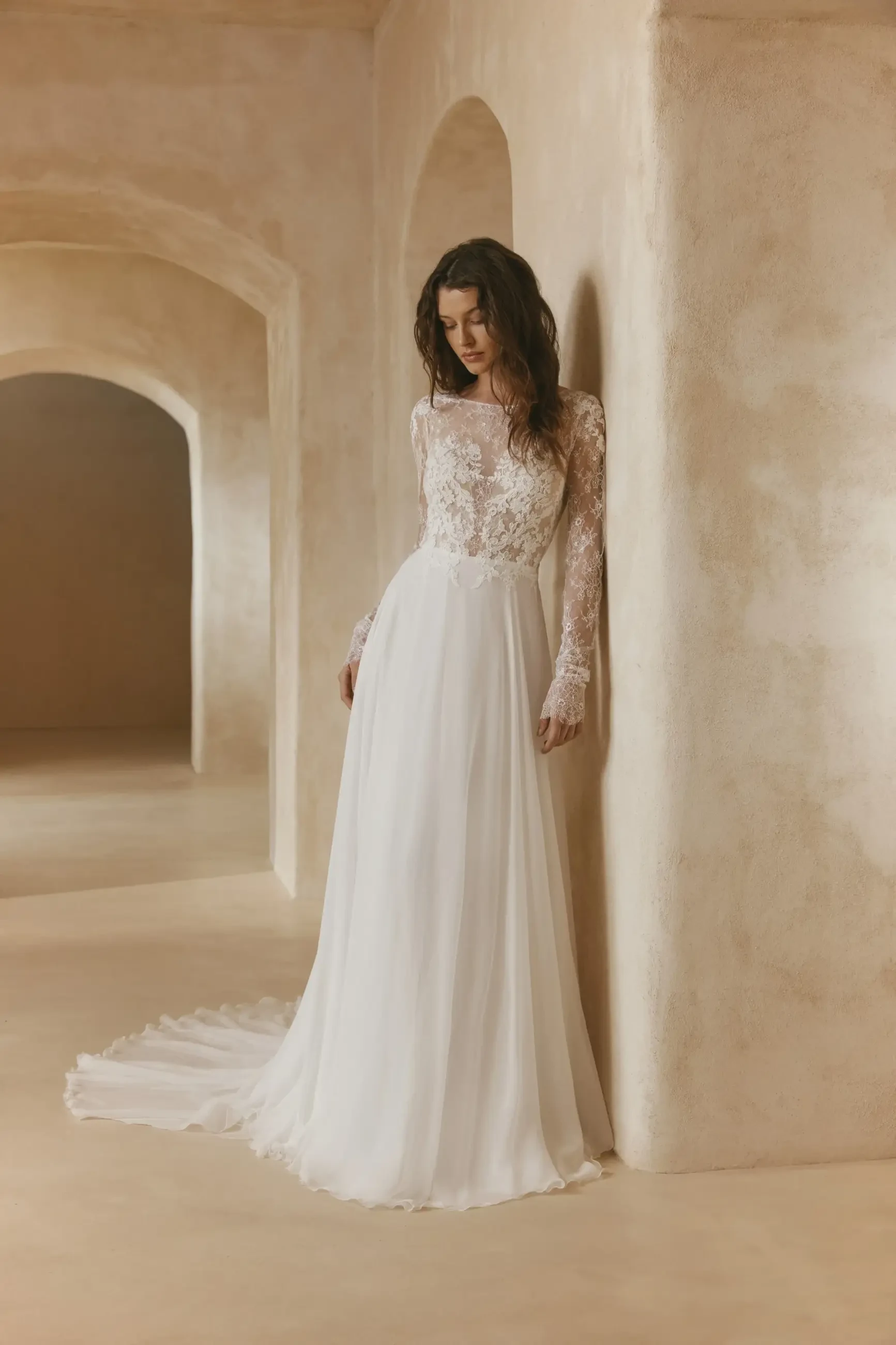 A bride wearing a long-sleeved, lace wedding dress stands against a textured wall in an elegantly simple setting.