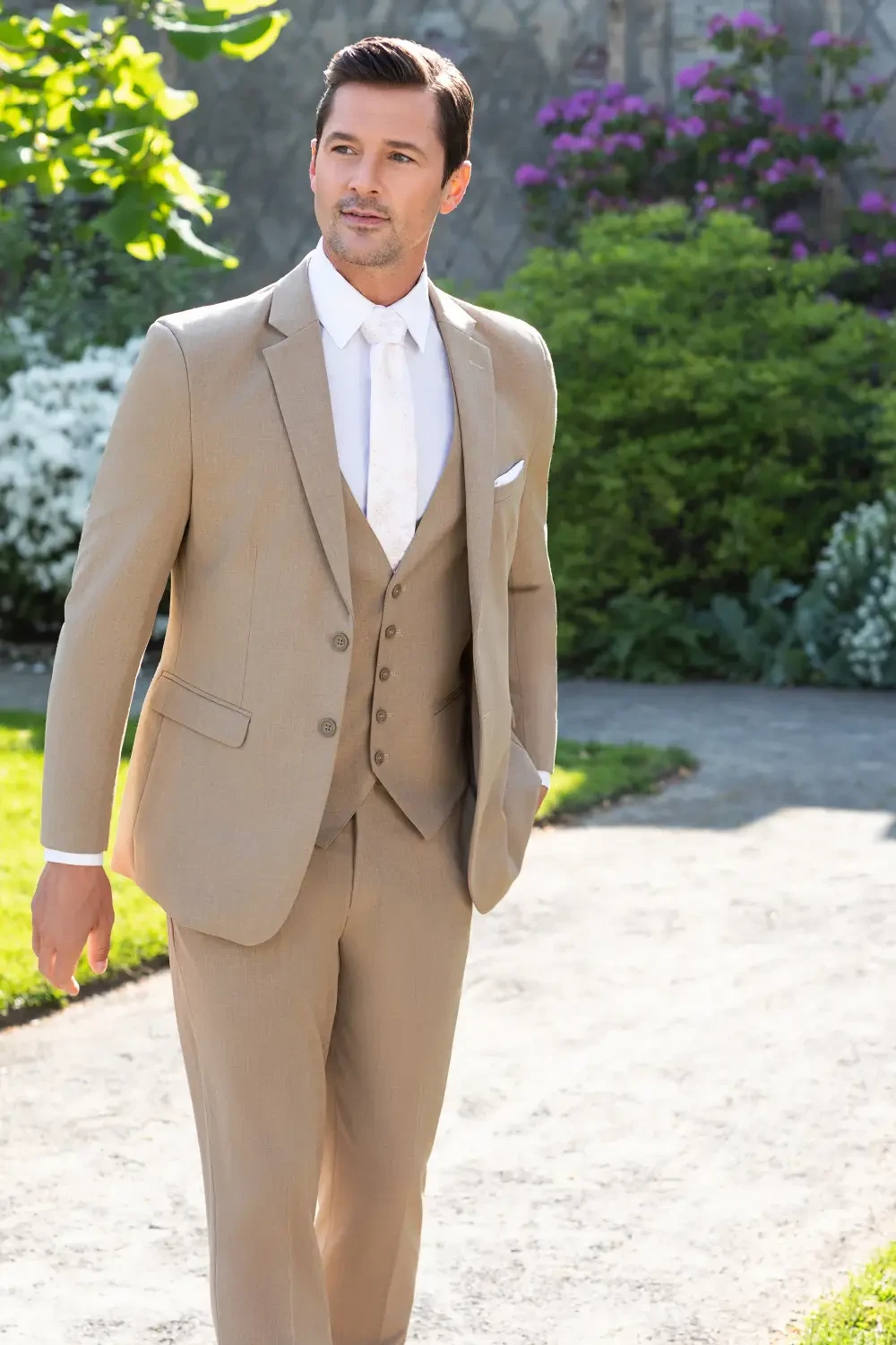 A man in a stylish beige suit with a white shirt walks down a path surrounded by greenery and flowers.