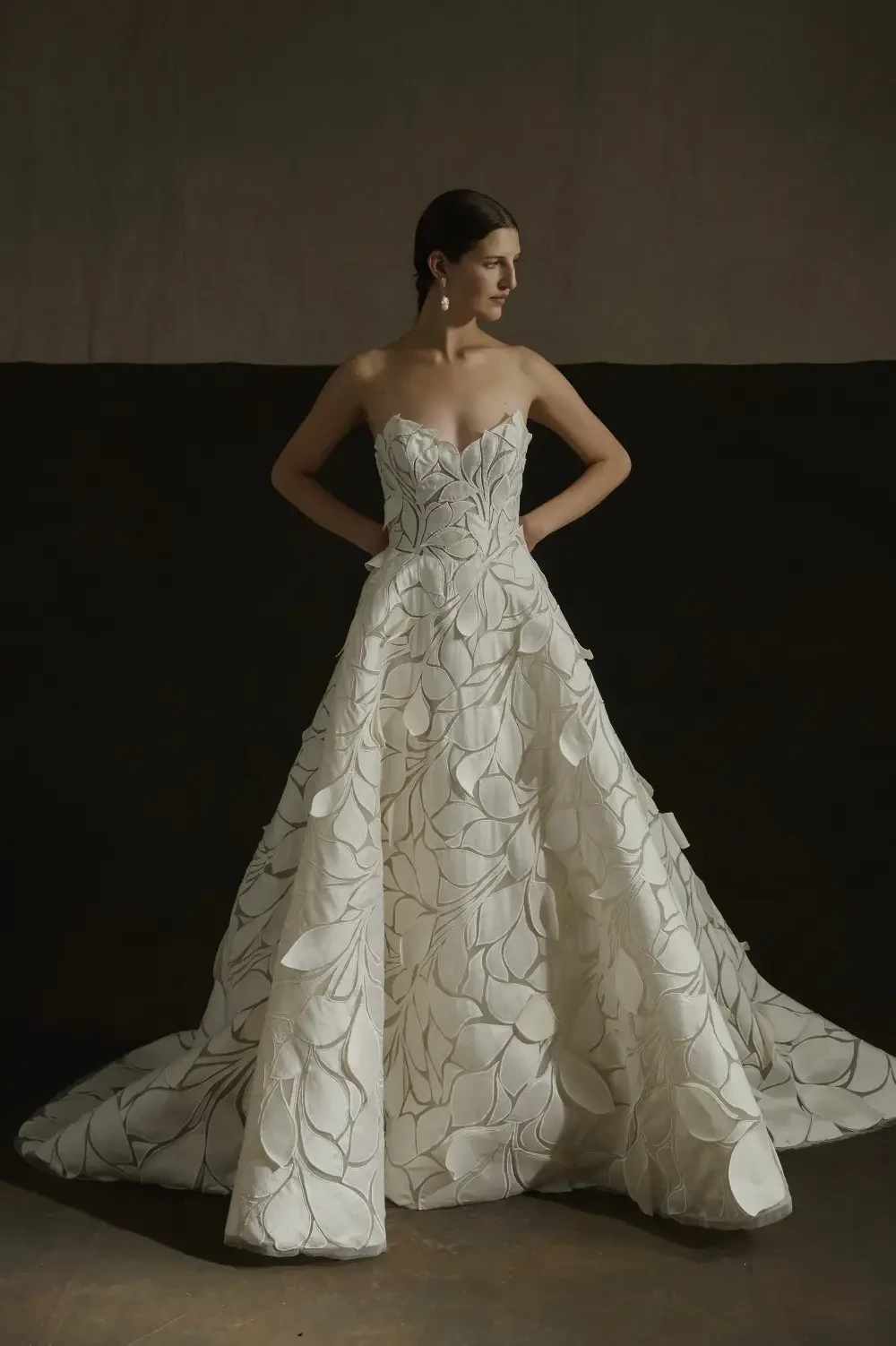 A woman in a strapless, intricately designed white gown with floral patterns, standing elegantly with her hands on her hips against a dark background.