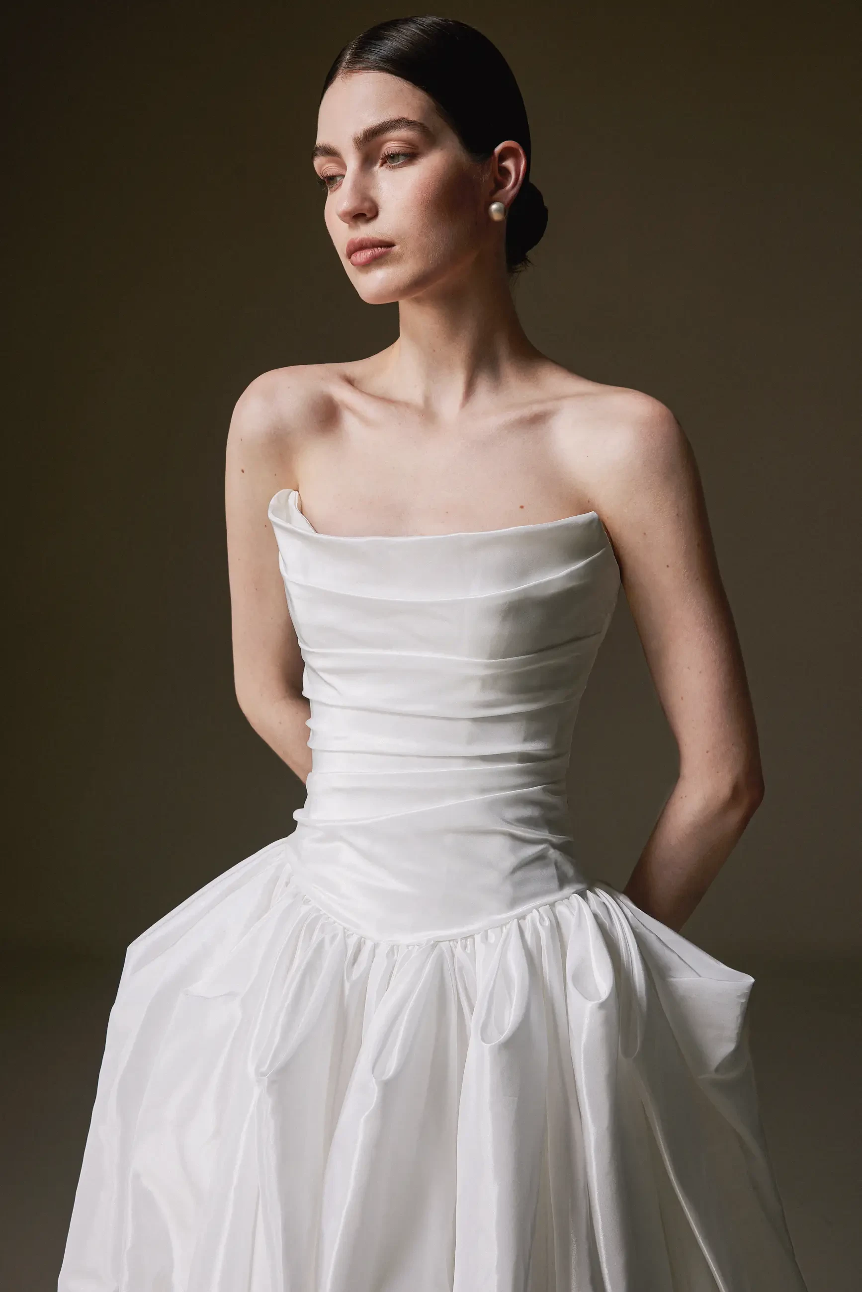 A woman in an elegant white, sleeveless gown with a ruched bodice stands with her arms behind her back. She looks poised, exuding a serene confidence.