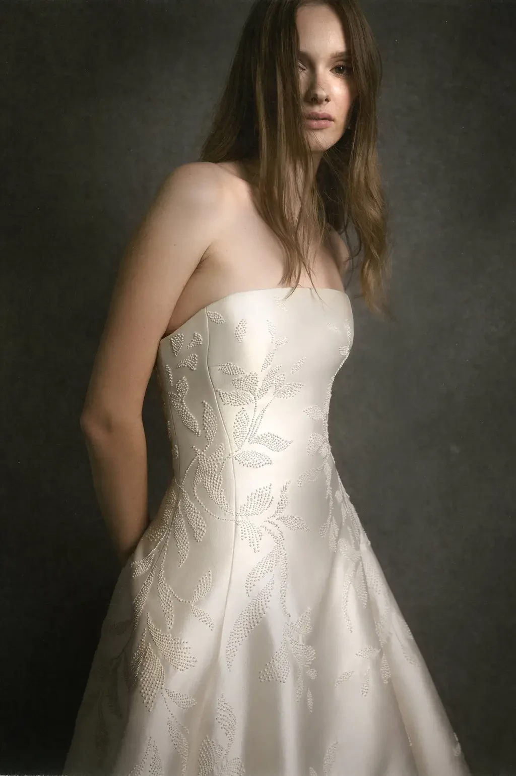 A woman wearing a strapless wedding dress with floral embroidery, standing against a dark background.