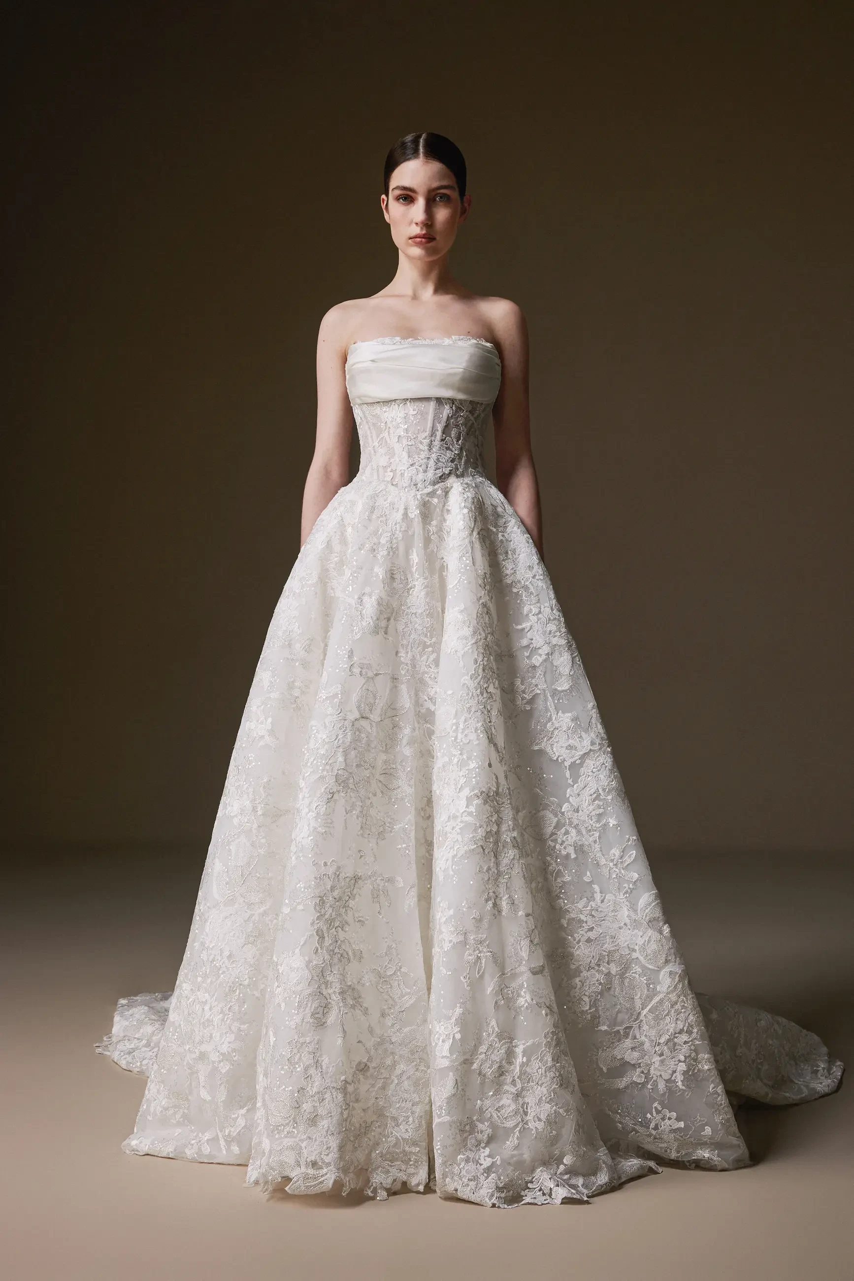 A woman in a strapless, lace wedding gown stands confidently against a neutral background. The dress is elegant with intricate floral patterns.