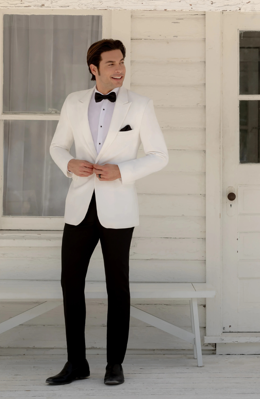 A man in a white tuxedo jacket and black pants standing outside a building, adjusting his jacket with a smile.