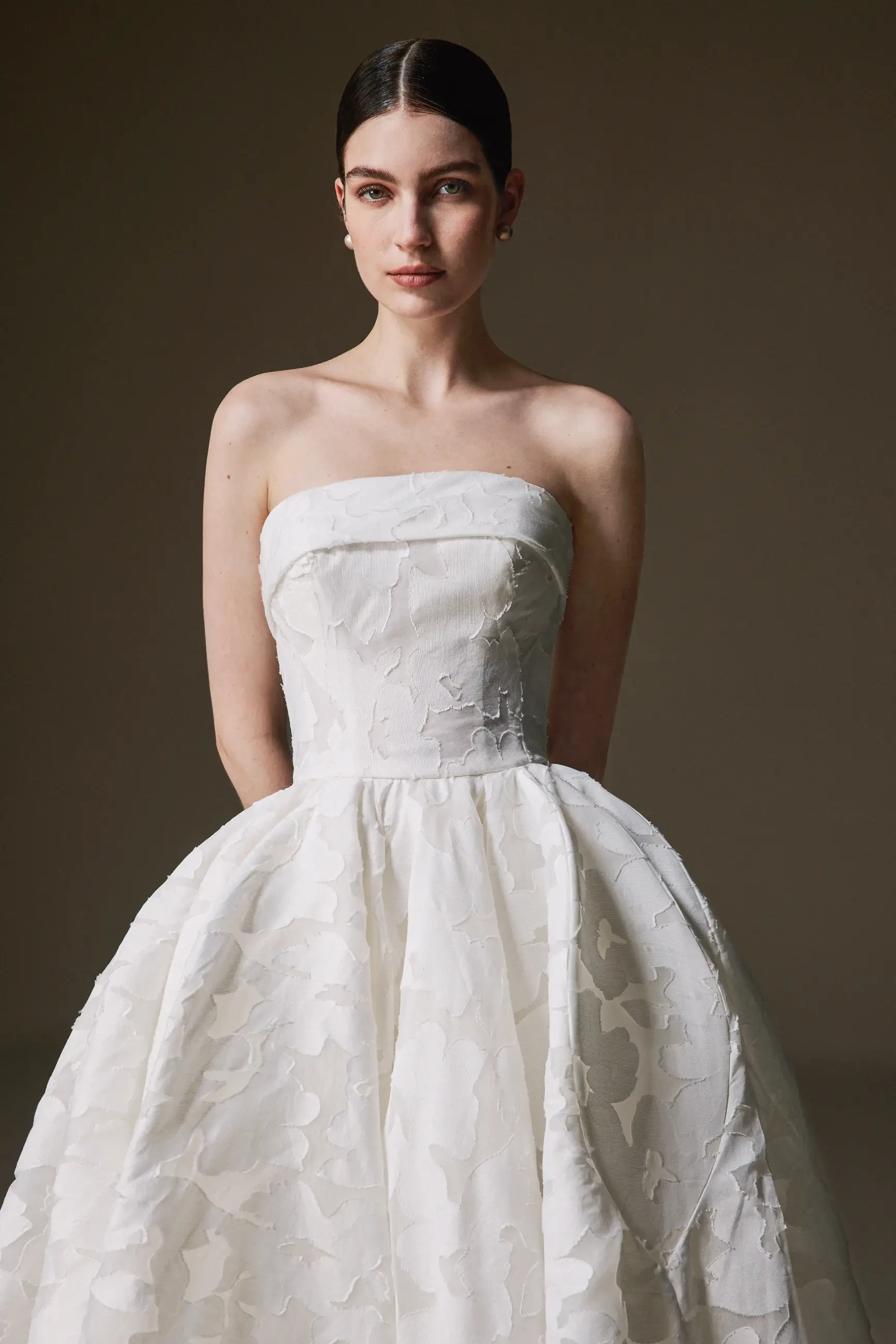 A woman in an elegant, strapless white wedding dress with a floral pattern stands against a neutral background, exuding a serene, poised demeanor.