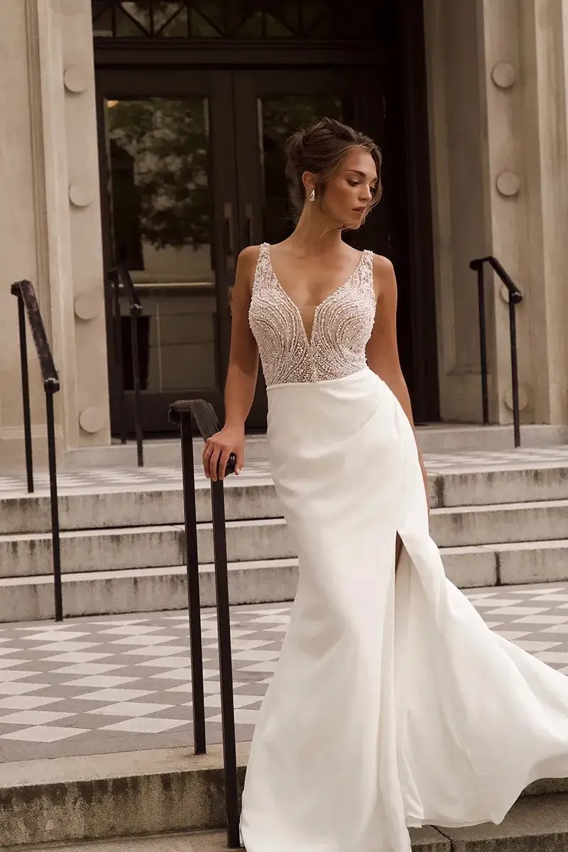 Model is wearing a crepe column wedding dress with a deep v neckline and pearl beading