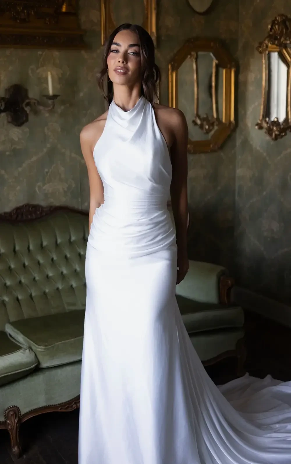 A woman in a fitted white gown stands in front of a vintage green sofa with ornate mirrors in a stylish interior.