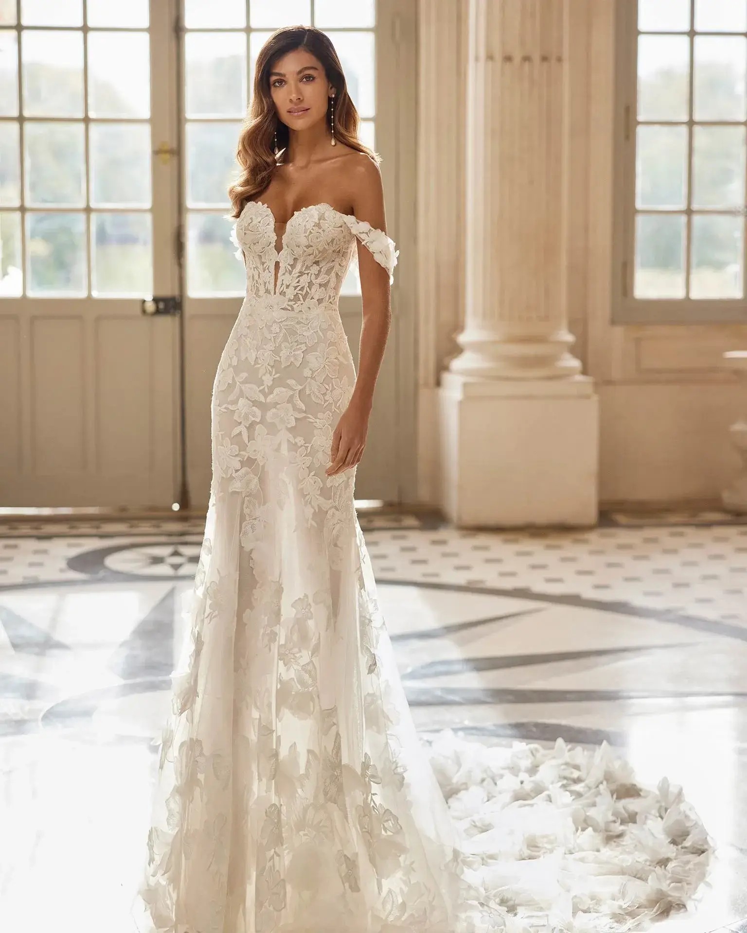 Model is wearing a fit and flare wedding dress with allover lace, off the shoulder sleeves, and a plunging neckline