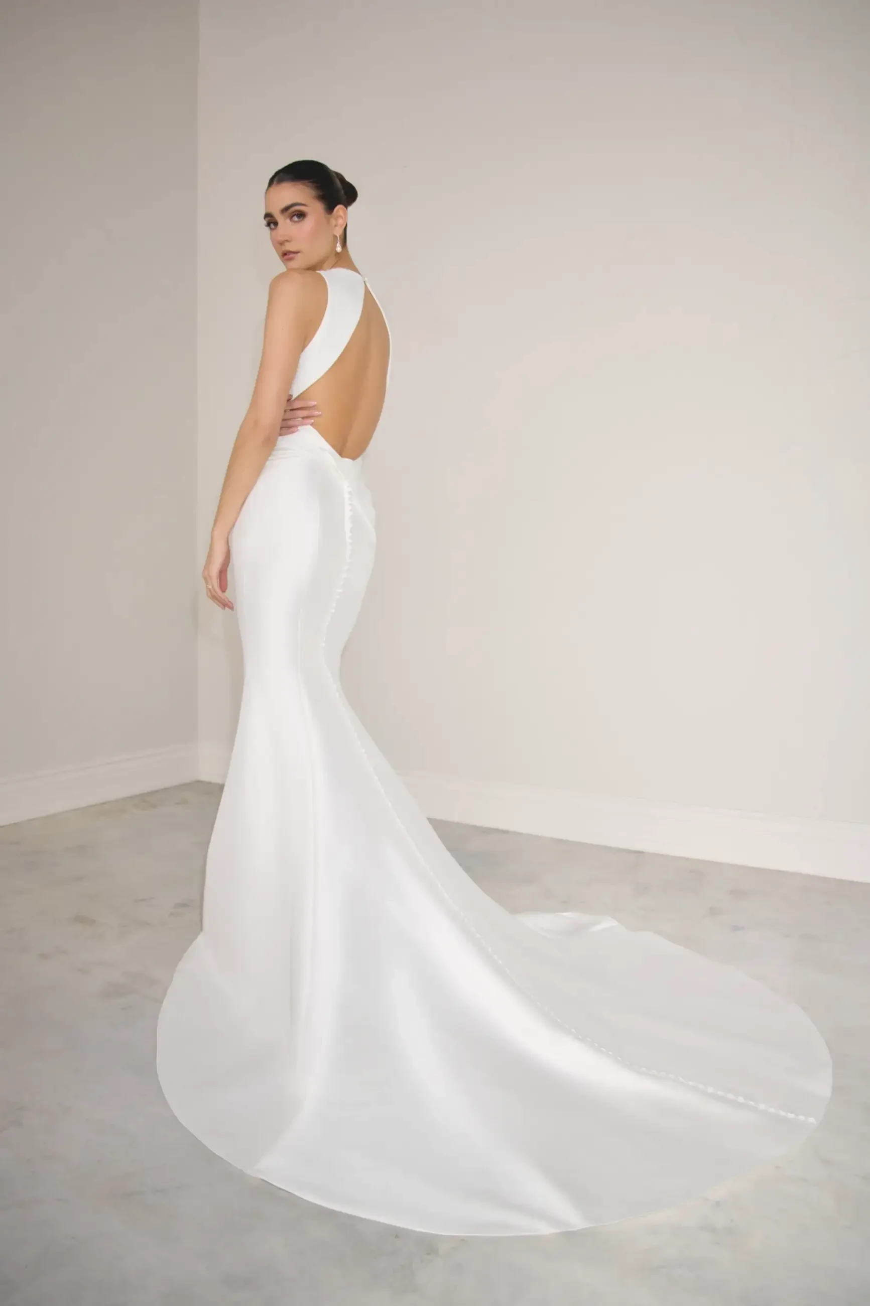 A model wearing a sleek, elegant white gown with an open back and a flowing train, posing against a neutral background.