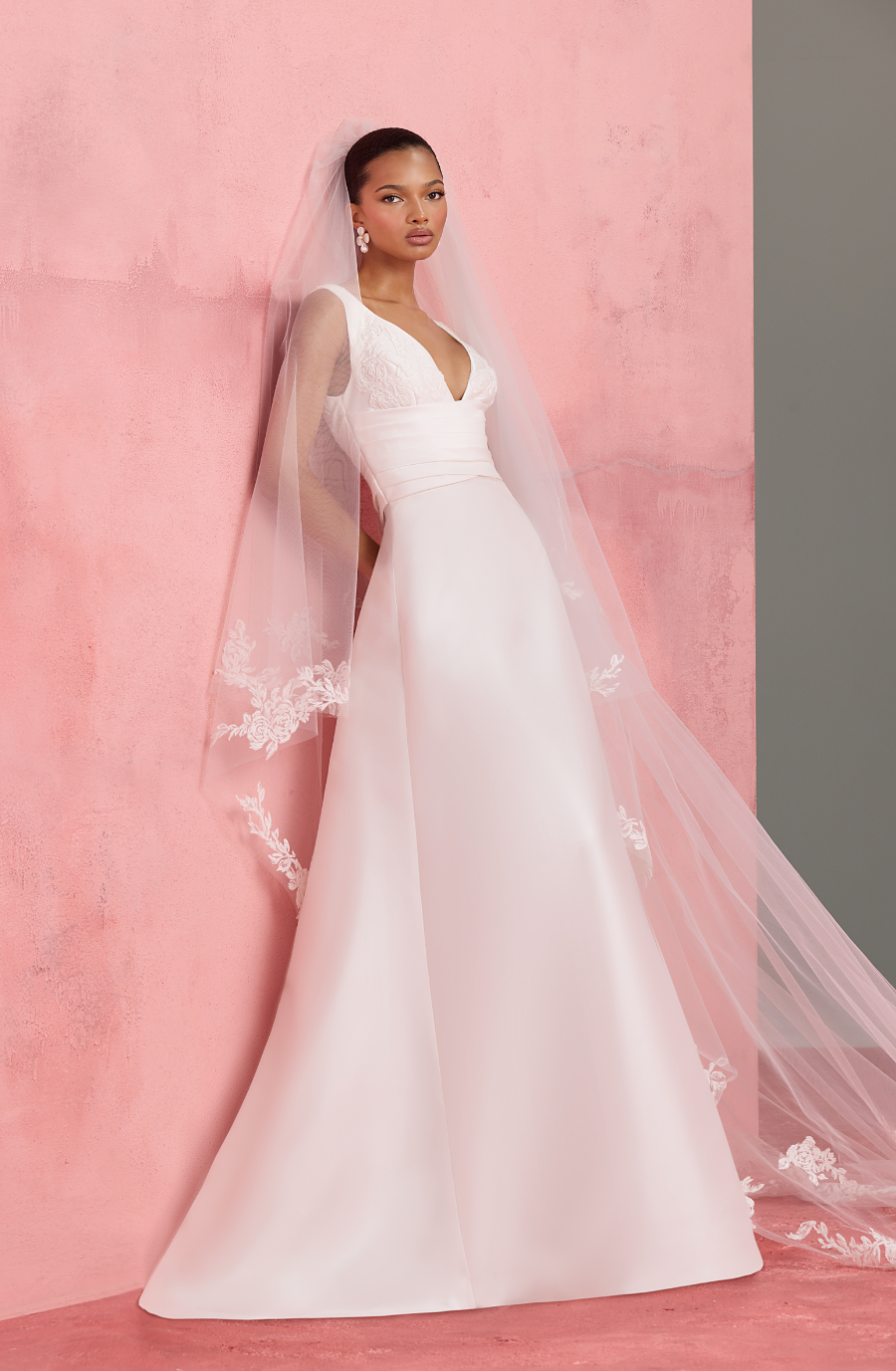 A bride in an elegant white wedding dress poses against a pink textured wall. The gown has a deep V-neck and delicate floral lace veil. The tone is romantic and serene.
