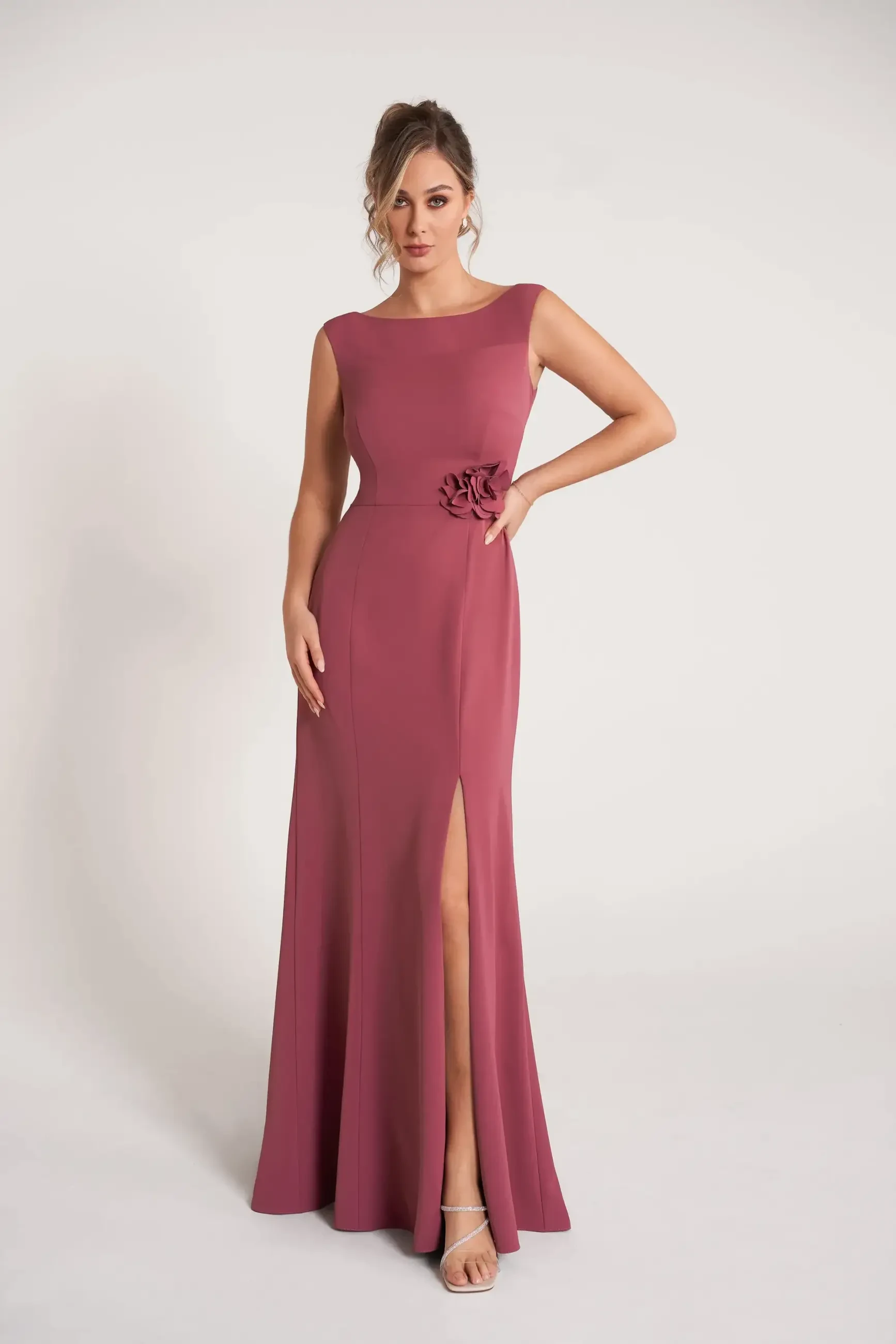 A woman wearing a fitted, sleeveless pink dress with a side slit and a floral accent on the waist, posing against a plain background.