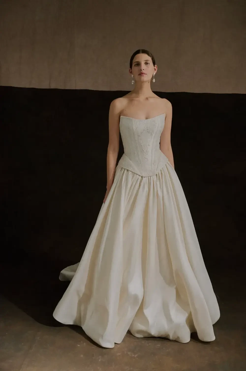 A model stands in a strapless white wedding gown, featuring a fitted bodice and a full skirt. The background is a neutral tone, enhancing the elegance of the dress.