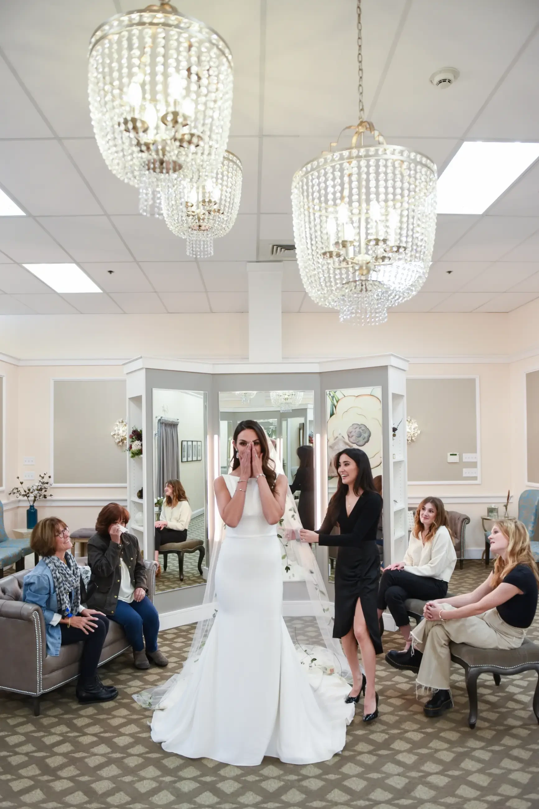 A bride in a white gown stands emotionally in a warmly-lit bridal shop, surrounded by supportive friends seated on couches. Elegant chandeliers hang overhead.