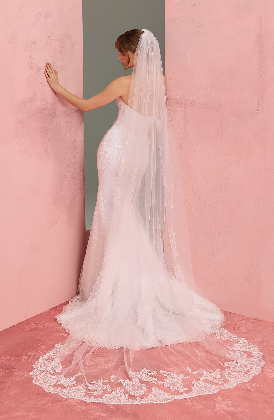 A bride in a flowing white dress and veil stands between pink walls, touching one. The veil elegantly trails on the floor, creating a delicate lace circle.