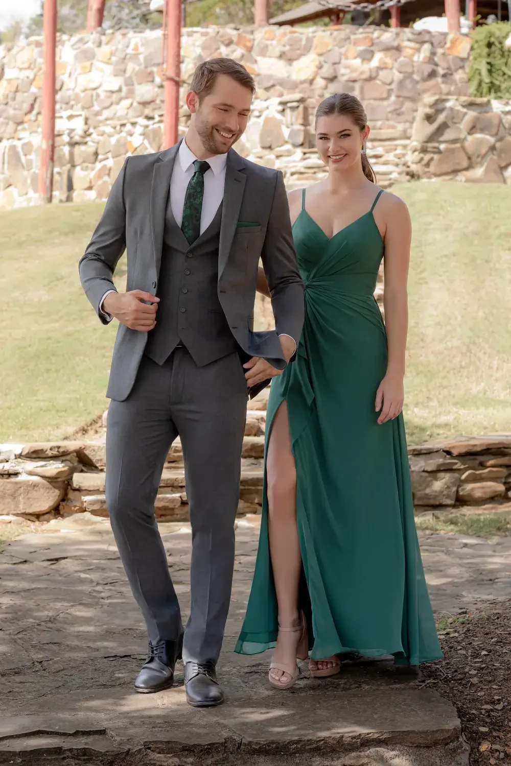 A man in a gray suit and a woman in a green dress with a thigh-high slit are walking together outdoors, smiling.