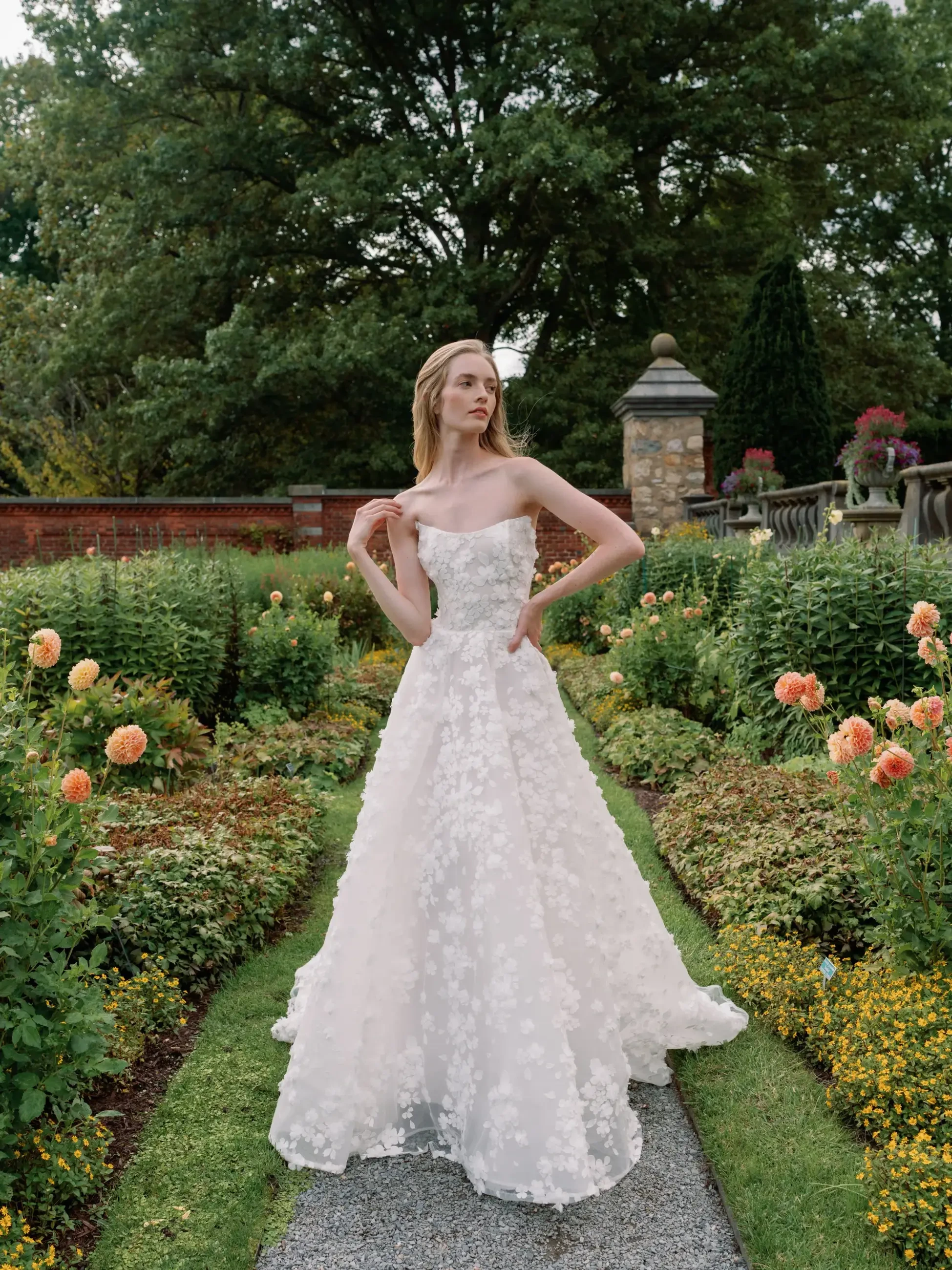 A model in a white floral gown poses in a lush garden with colorful flowers and greenery.