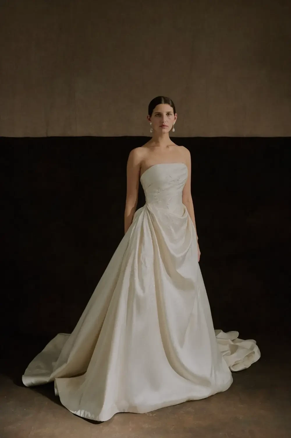 A woman in an elegant strapless wedding gown stands against a dark background.