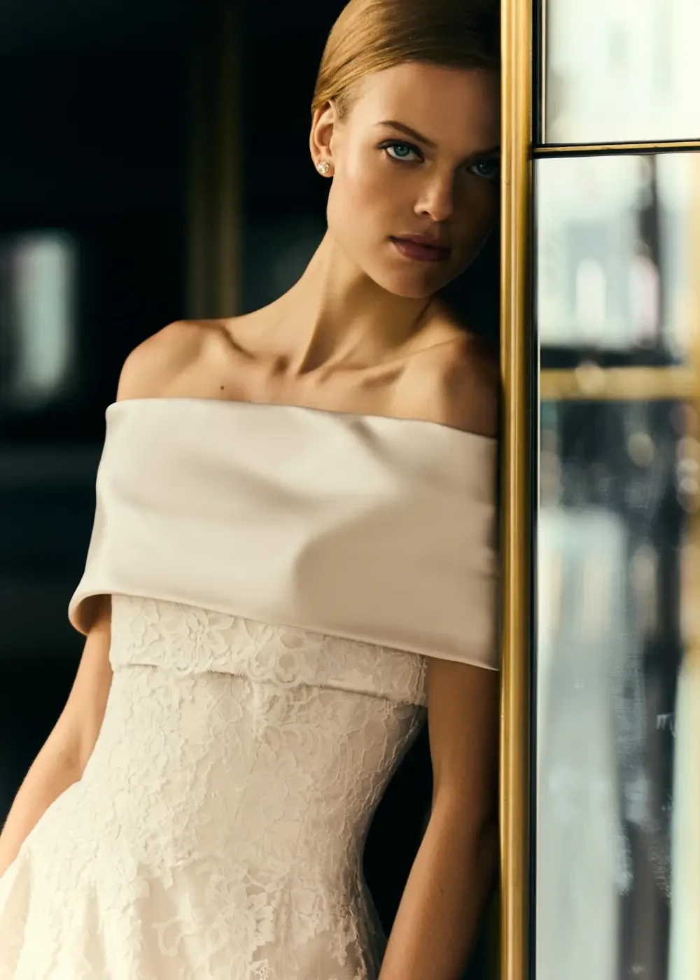 A model wearing a strapless, off-white dress with a lace overlay and a smooth satin bodice, standing close to a reflective surface.