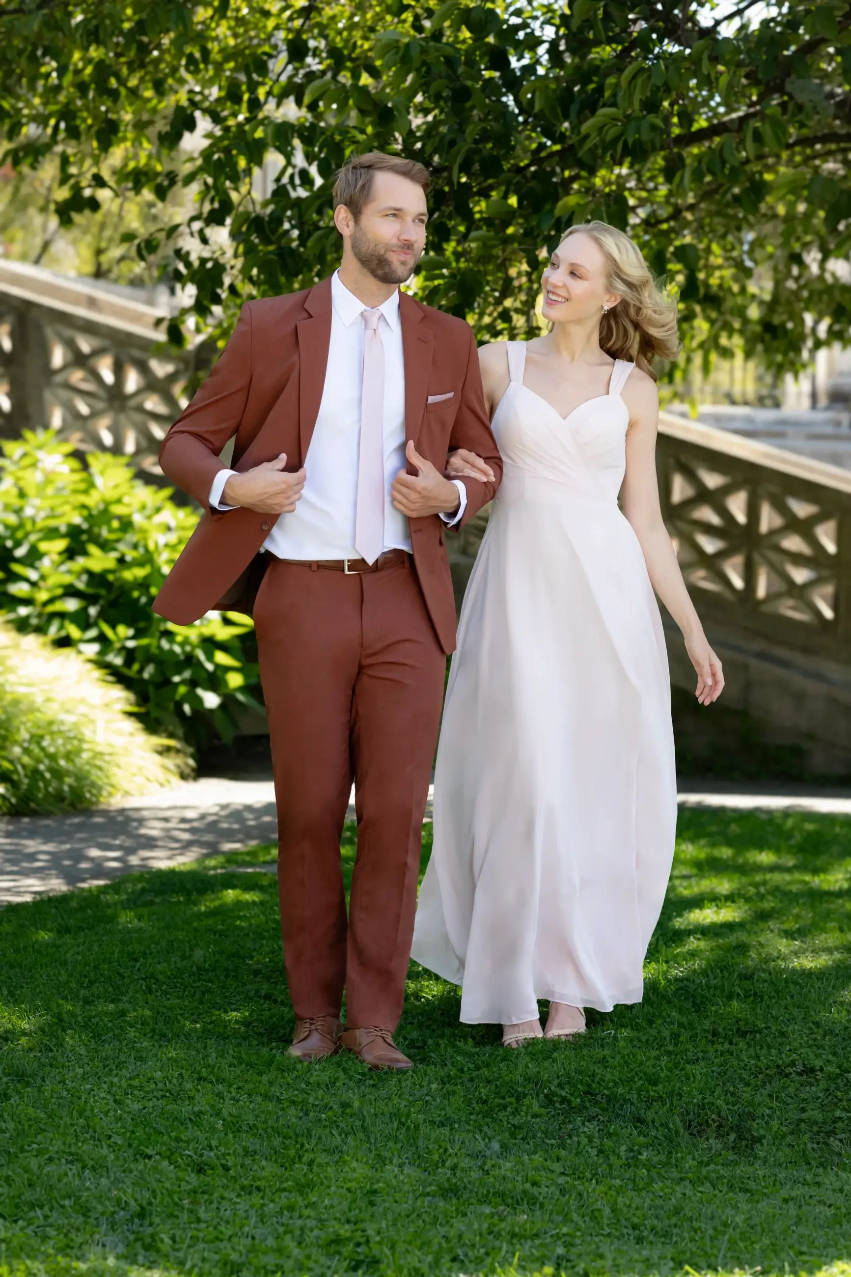 A man in a rust-colored suit and a woman in a light pink dress walk together on a grassy area with greenery in the background.