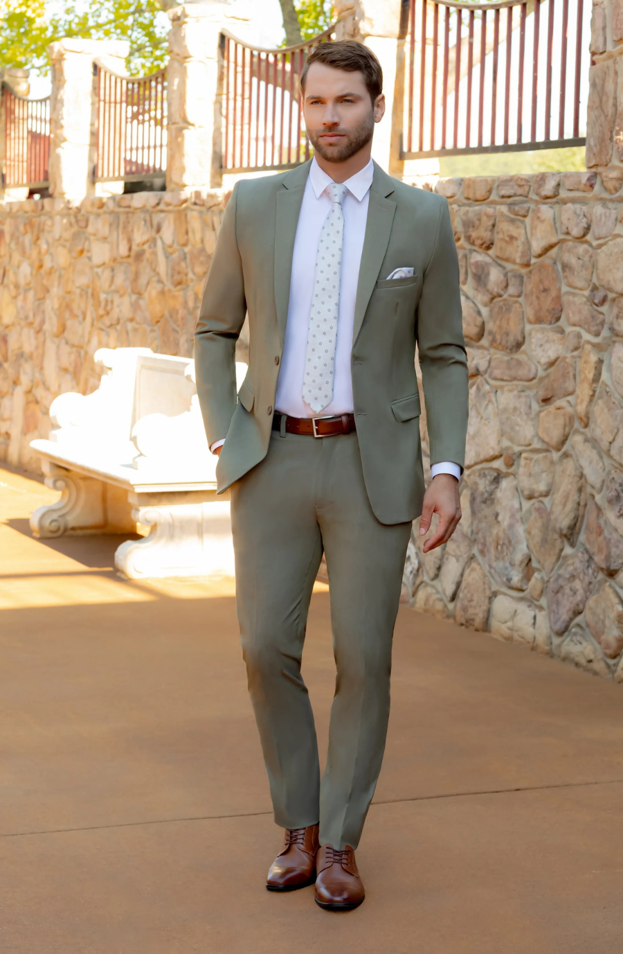 A man in a light green suit stands confidently outdoors, against a stone wall backdrop, wearing a white shirt and a patterned tie, with brown dress shoes.