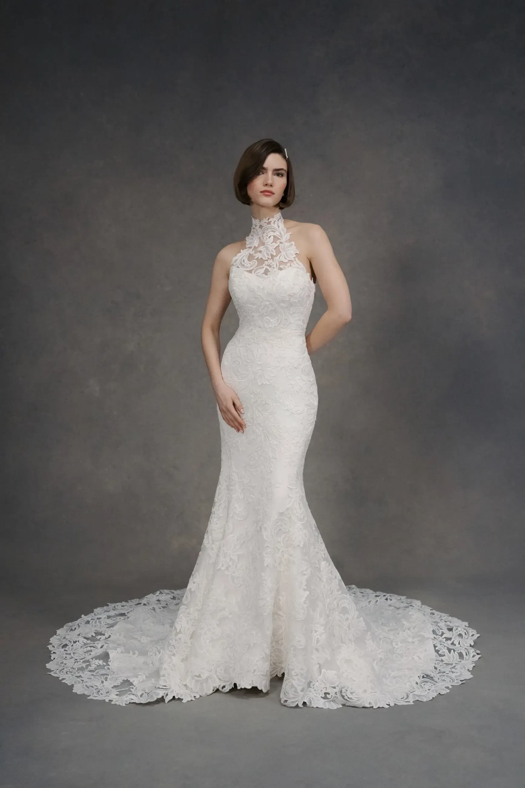 A model wearing an elegant white lace wedding dress with a fitted silhouette and a flared train, against a neutral background.