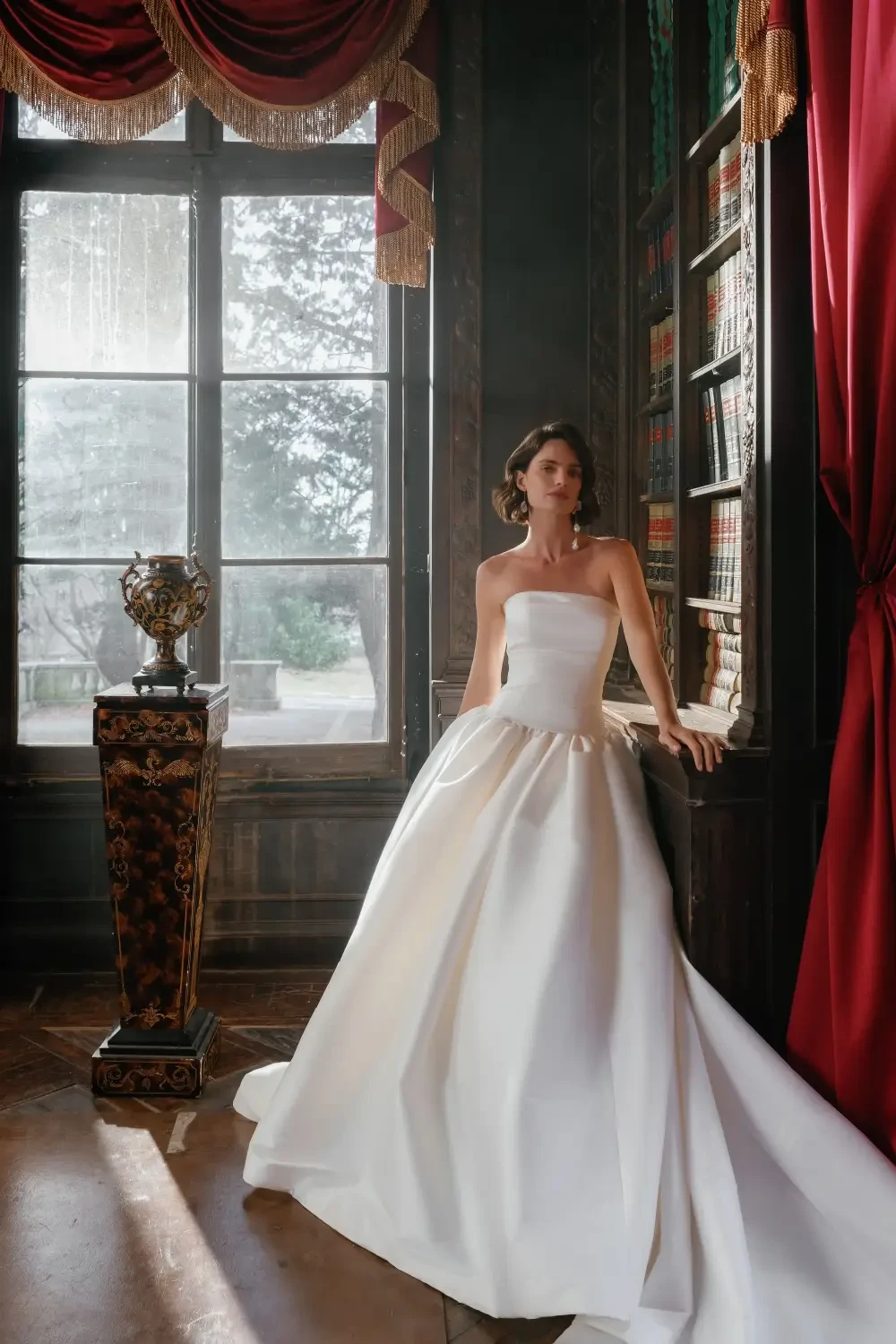 A woman in a elegant white strapless gown stands in a grand room with large windows, surrounded by bookshelves and luxurious curtains.