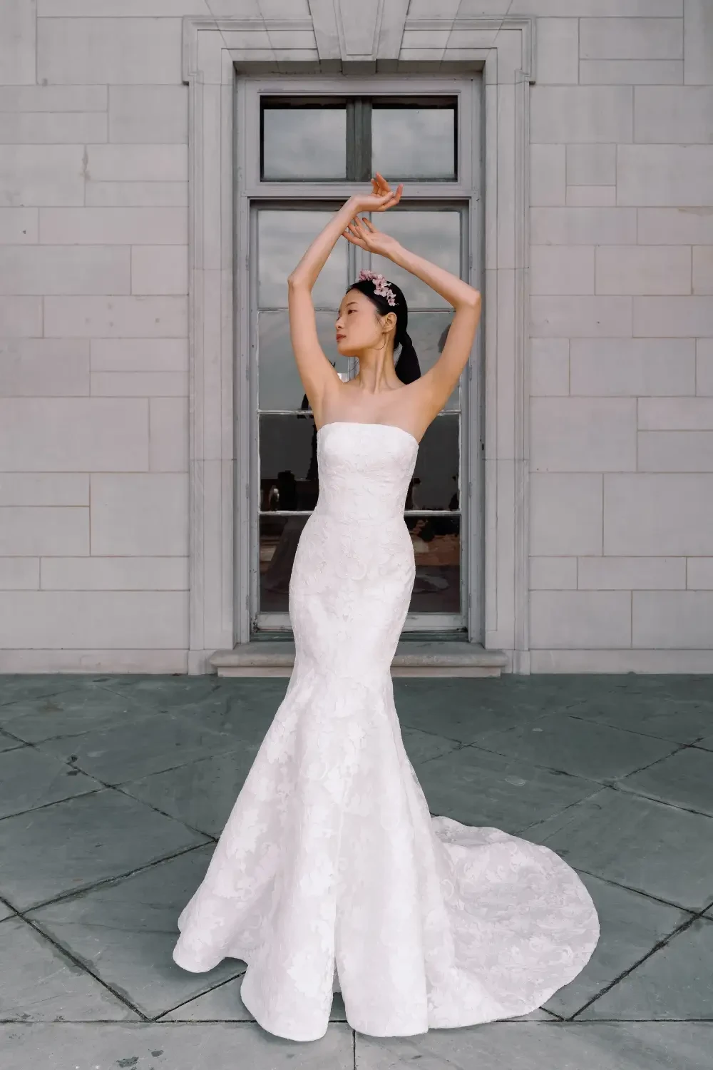 A woman wearing a strapless white wedding gown poses gracefully with her arms raised, standing outdoors in front of a large window.