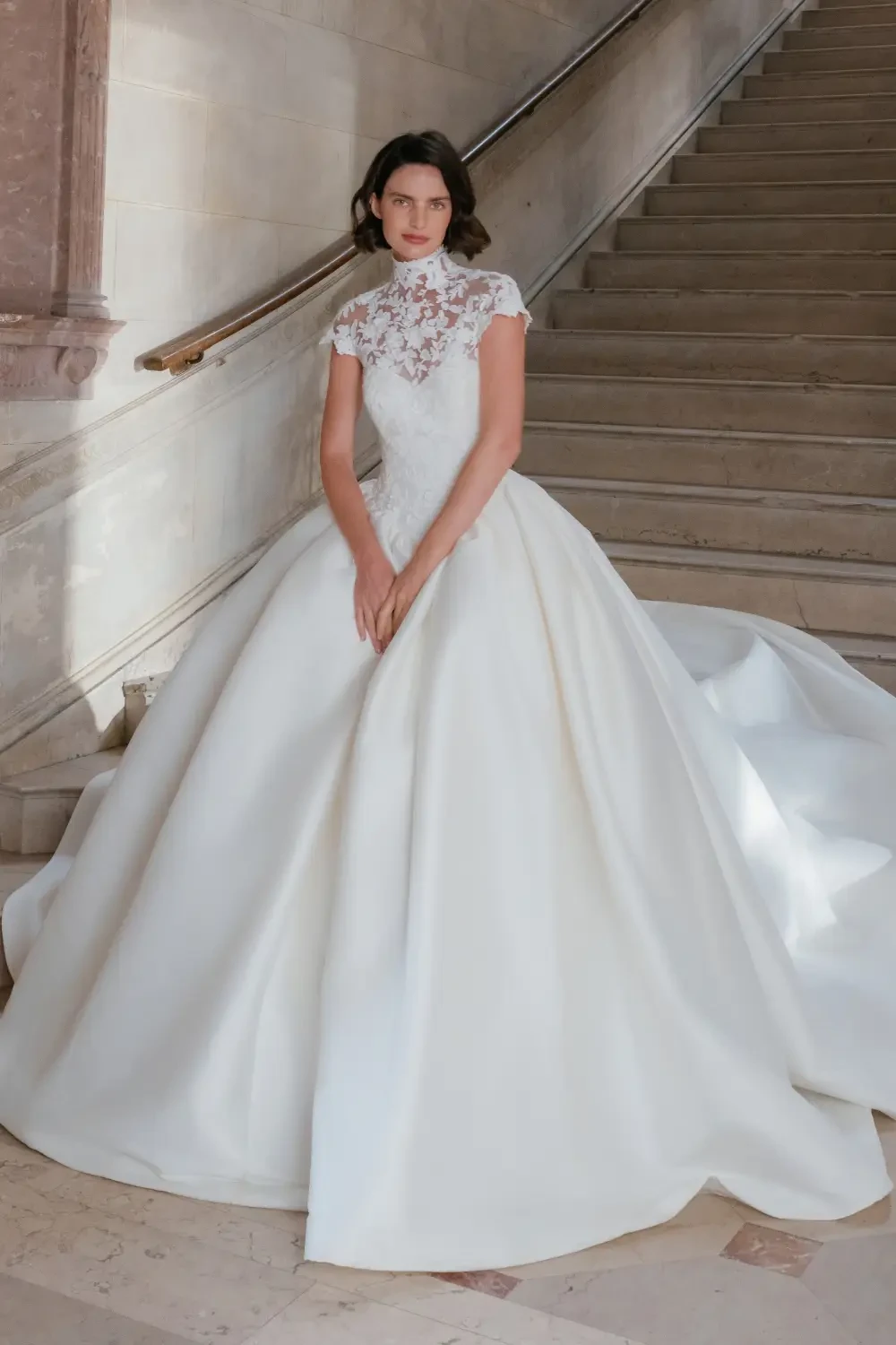 A model wearing an elegant white wedding dress with a lace bodice and a full skirt, posing on a staircase.