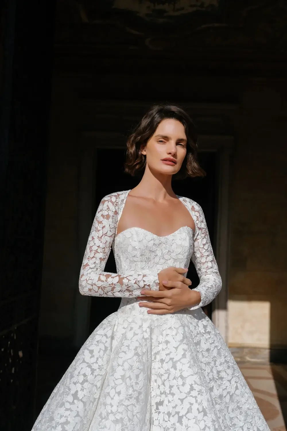 A woman in a white lace wedding dress stands elegantly, with a serene expression, in a well-lit environment.