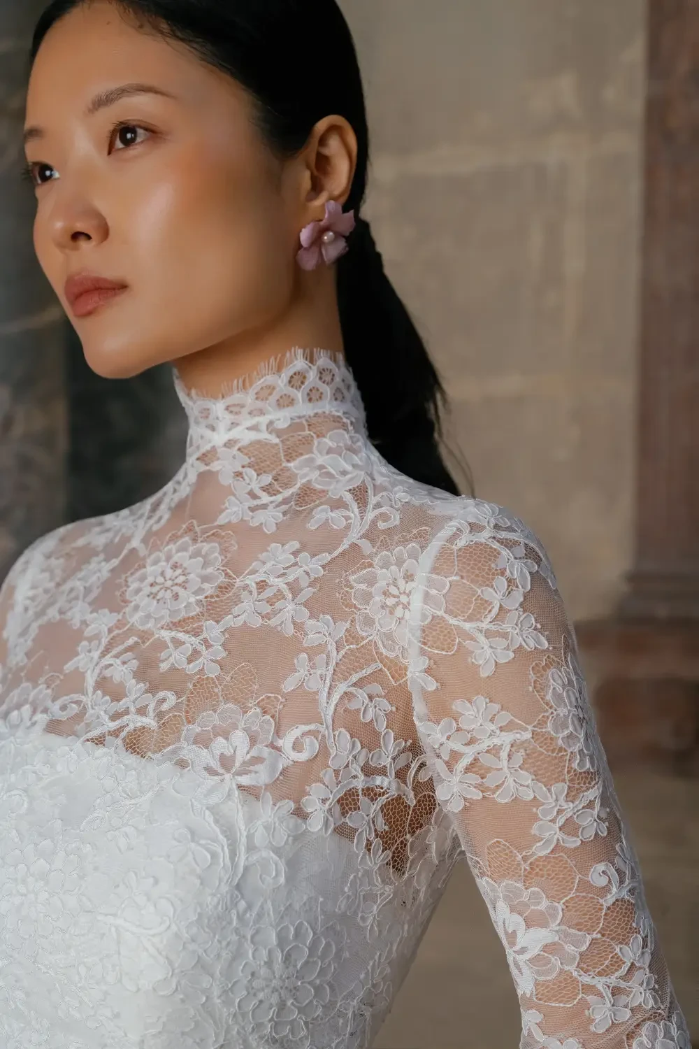 A woman wearing a white lace dress with a high neck and floral details, featuring a sleek hairstyle and earrings.